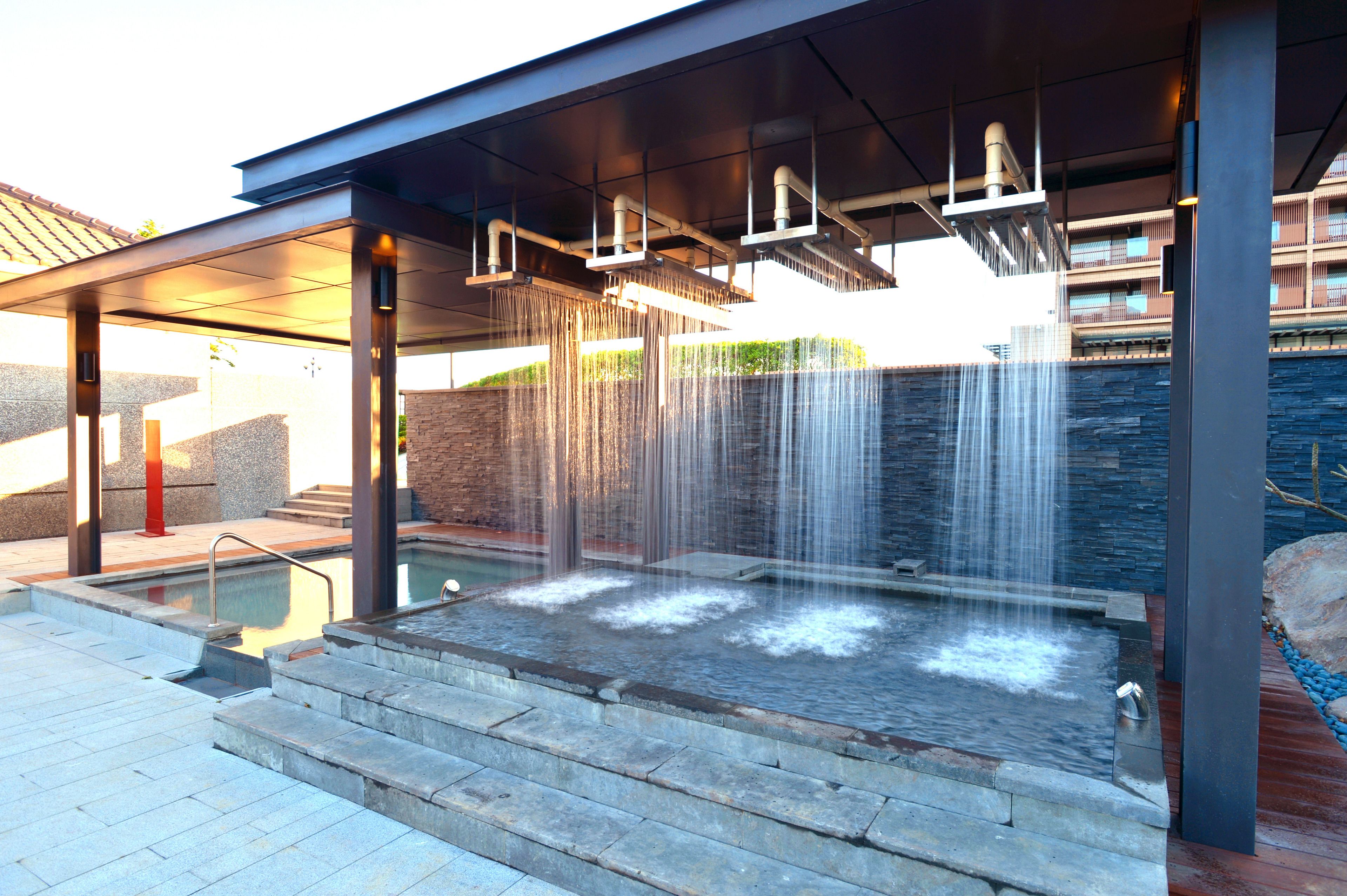 Un área de spa moderna al aire libre con hidromasaje, múltiples fuentes de agua y una zona de estar cubierta.