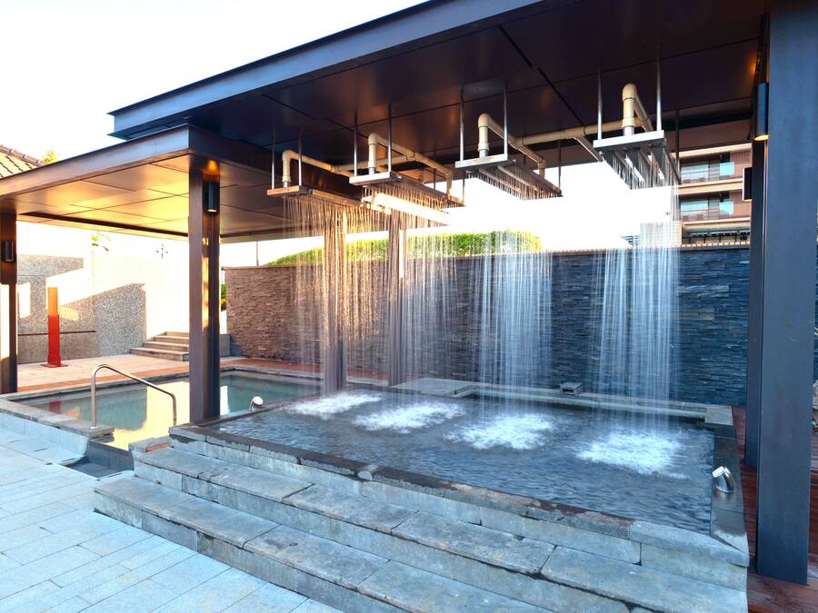 Un área de spa moderna al aire libre con hidromasaje, múltiples fuentes de agua y una zona de estar cubierta.