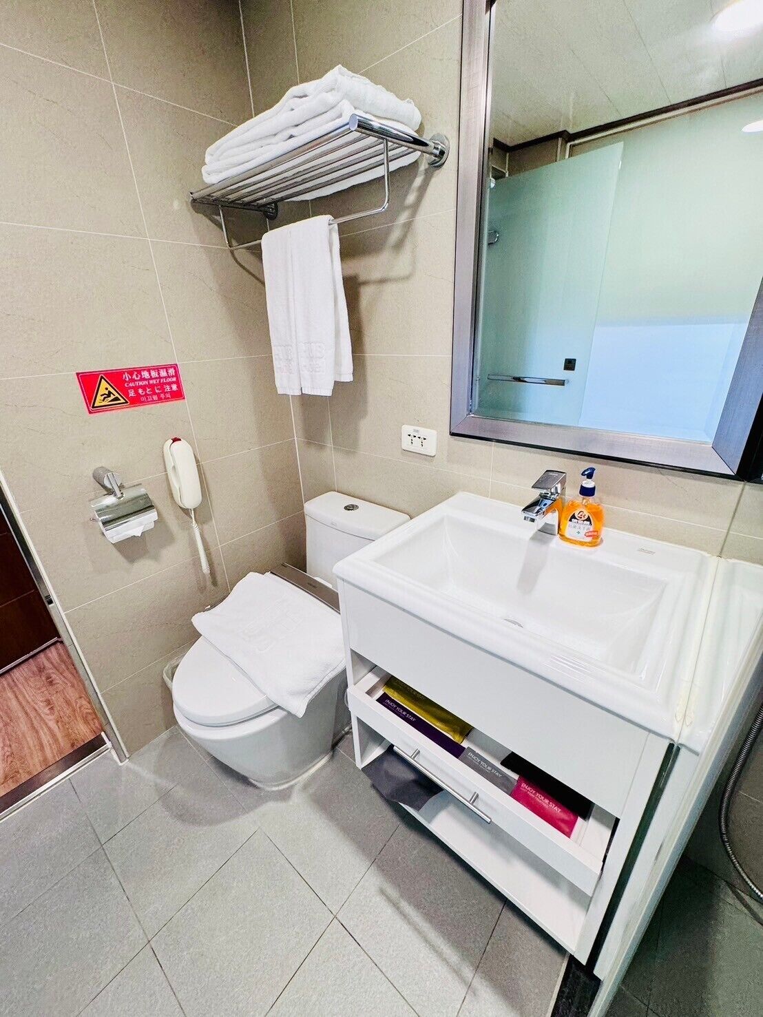 Baño con inodoro, lavabo y perchero. Hay un letrero de precaución en la pared y un teléfono en el suelo.