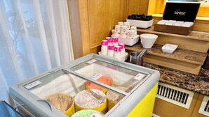Una exhibición de postres con diversos sabores de helado en una vitrina, rodeada por un mostrador con bandejas de galletas y una caja con el logo de la marca.
