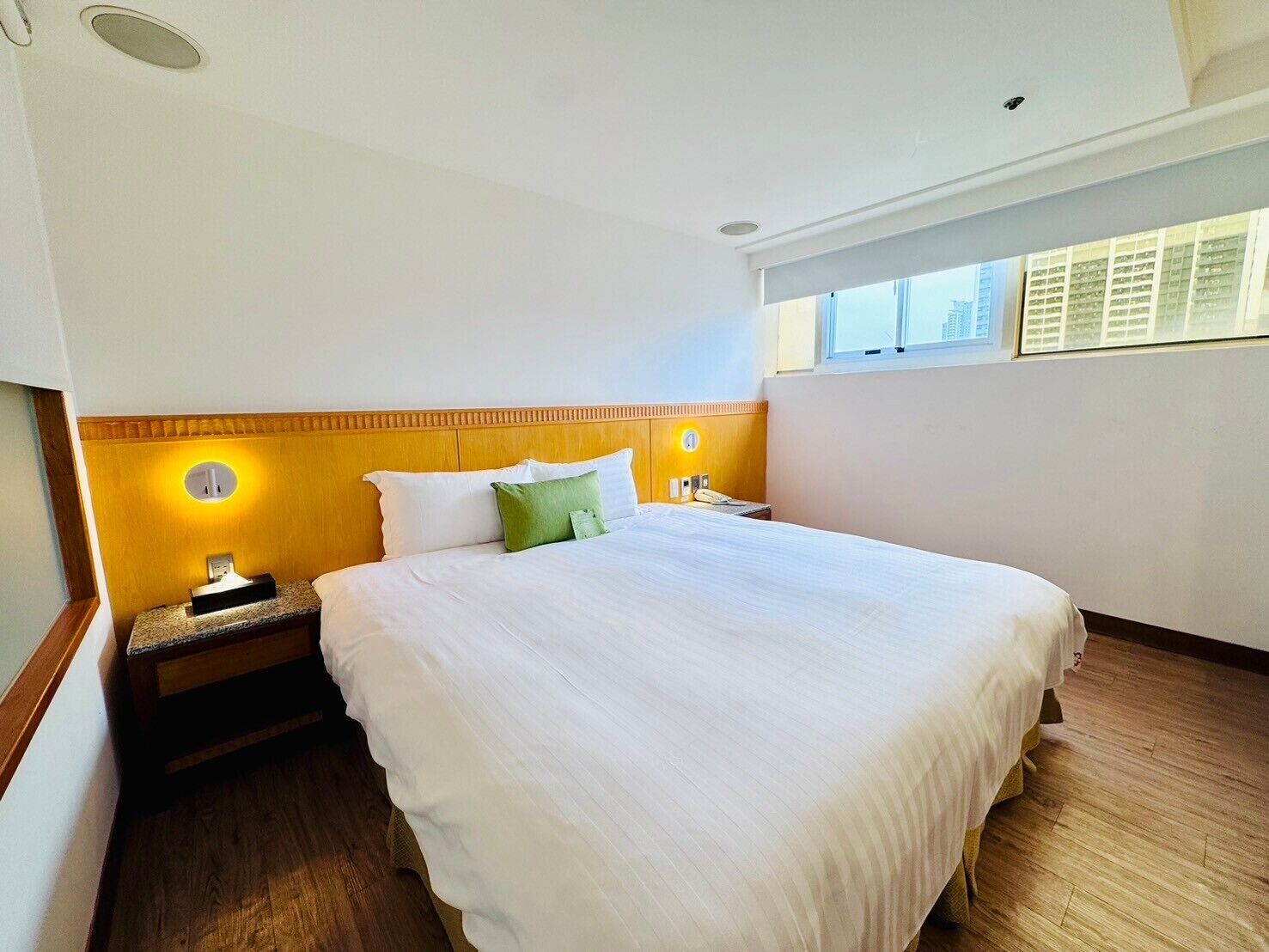 A hotel room with a large bed, wooden headboard, bedside lamps, and a window with a view of buildings.