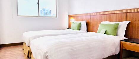 A hotel room with two beds, wooden headboard, a nightstand, and a window with a view of buildings.