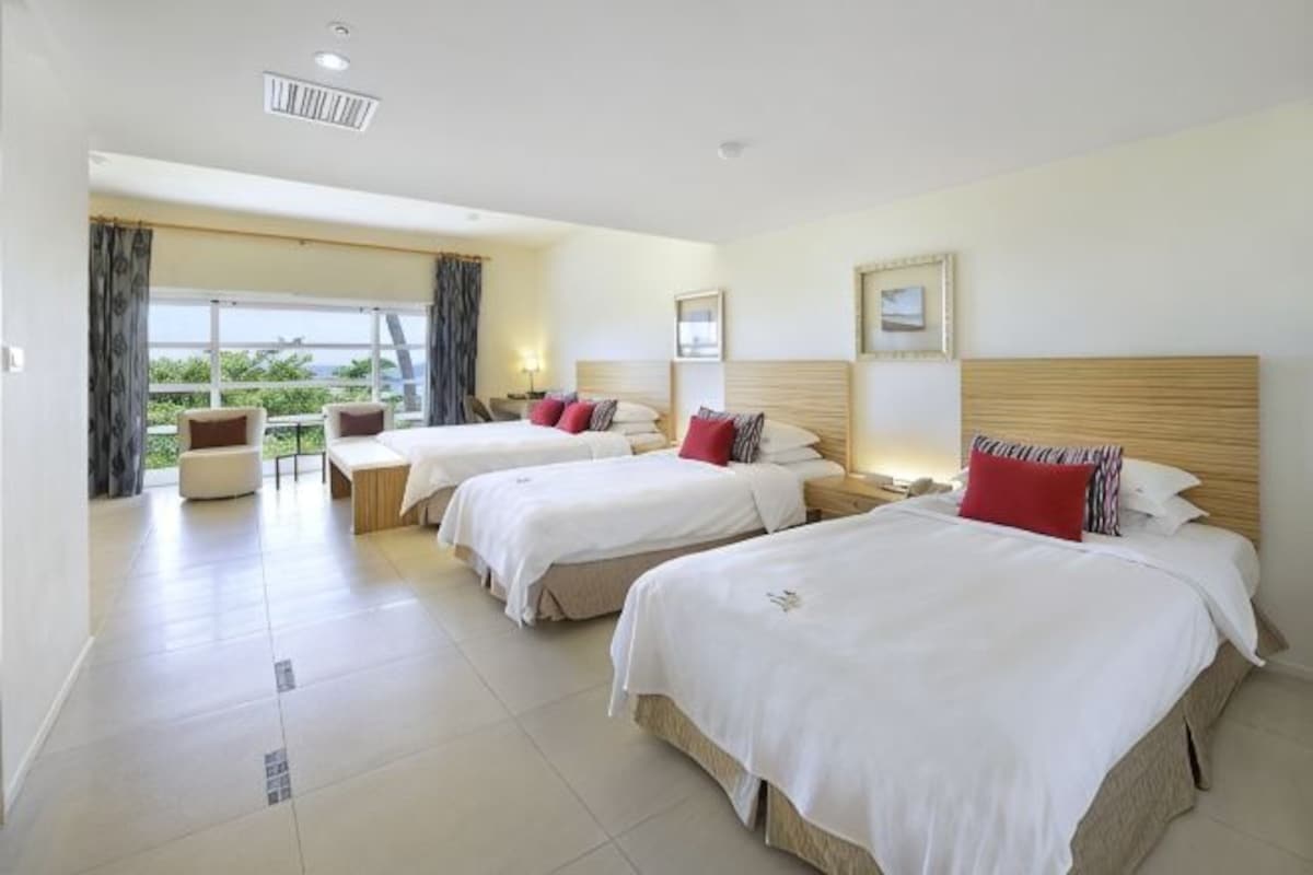 Habitación de hotel con tres camas, un espacio de descanso con mesa y sillas, y un gran ventanal con cortinas.