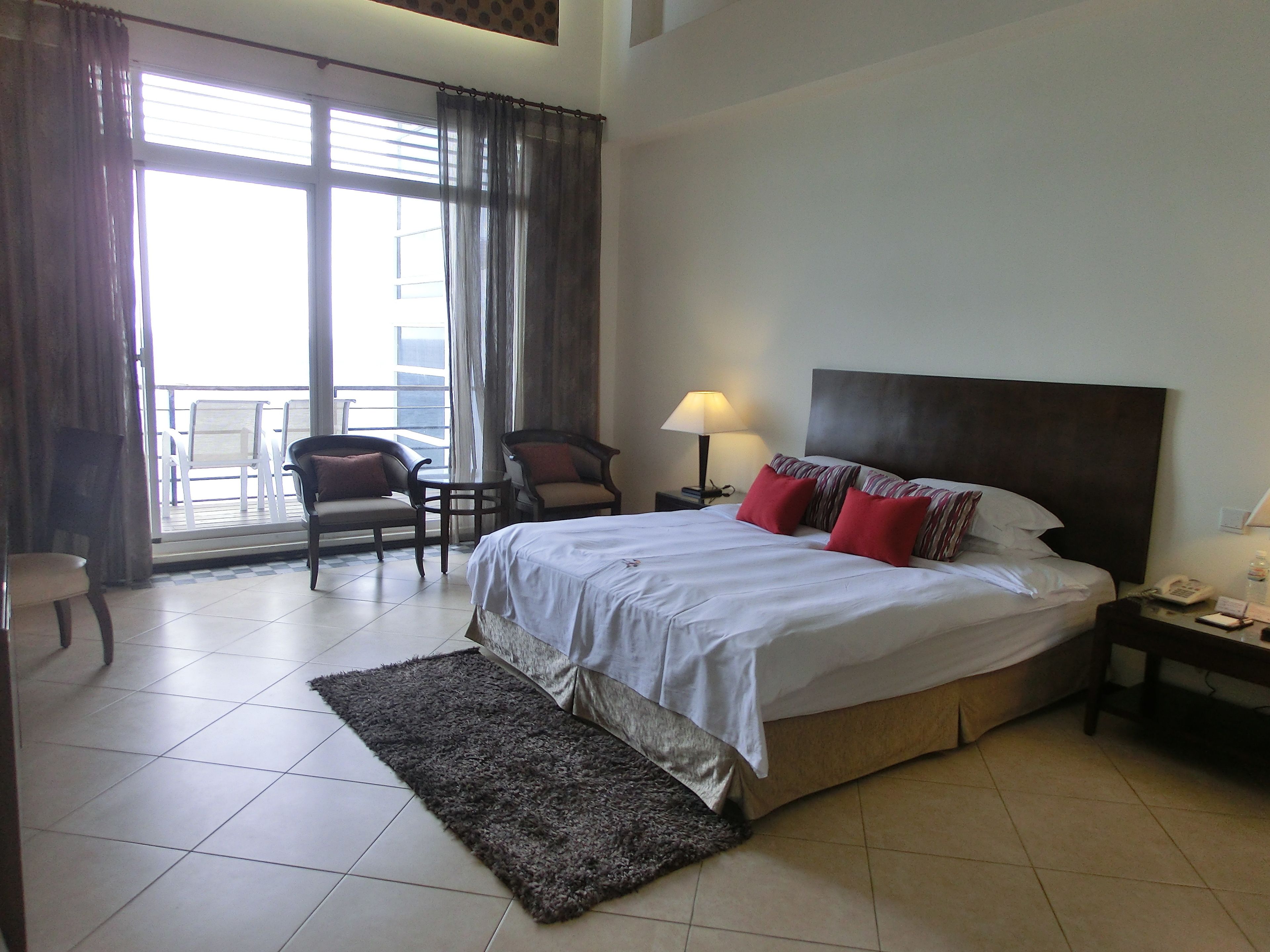 A hotel room with a bed, two chairs, a nightstand, a lamp, and a view of the outside.