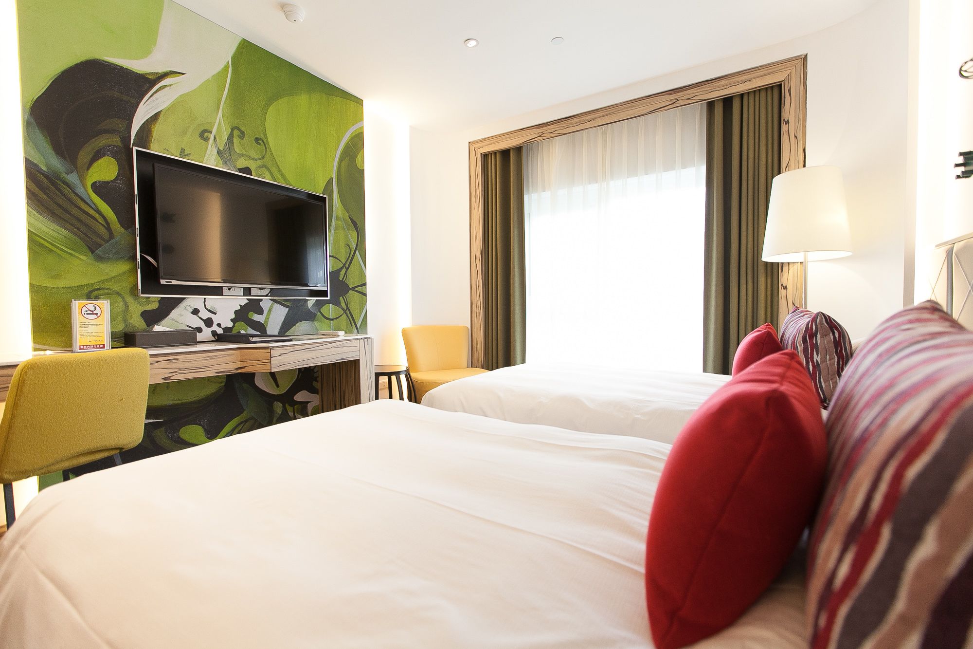 A hotel room with a bed, a desk with a television, a chair, and a window with curtains.