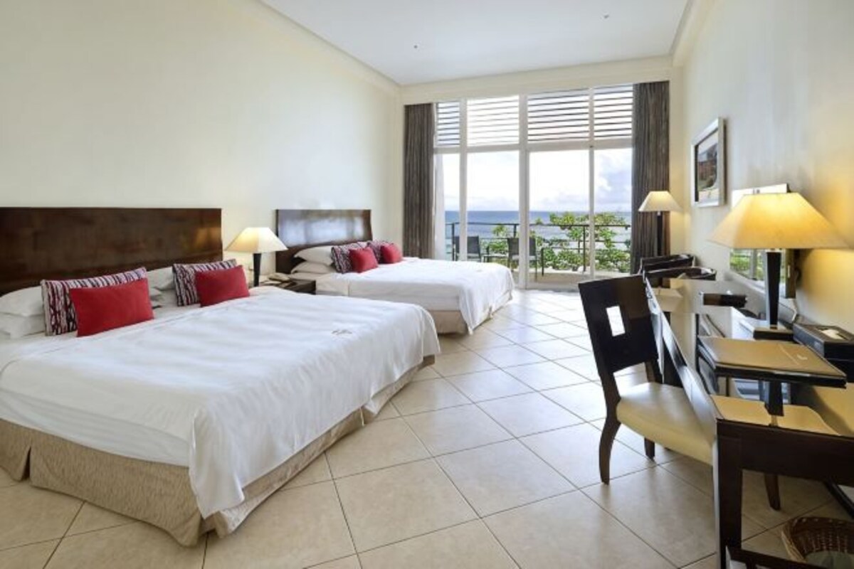 A hotel room with two beds, a dining table, chairs, and a view of the outdoors.