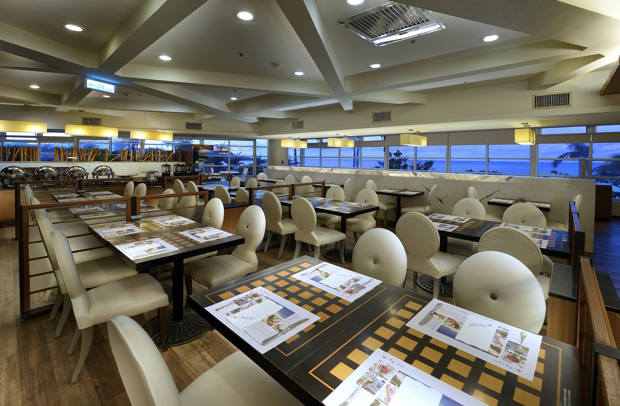 A restaurant with multiple tables and chairs, a view of the ocean, and a ceiling with recessed lighting.