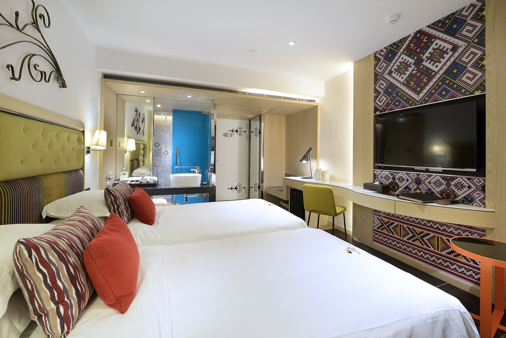Habitación de hotel con dos camas, un escritorio, una silla, un televisor y un baño visible a través de una puerta abierta.