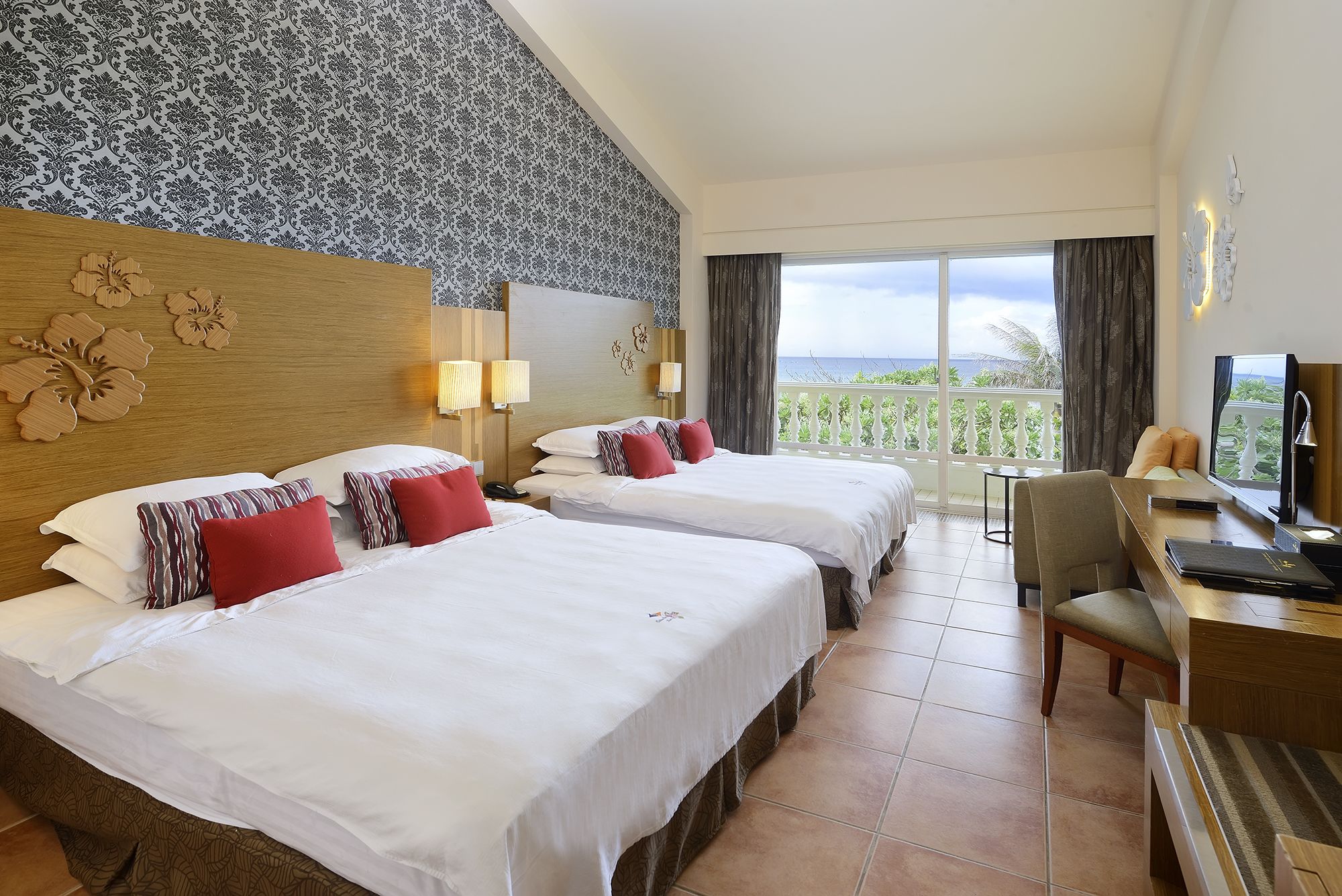 A hotel room with two beds, a desk, a chair, and a TV. The room has a balcony with a view of the ocean.