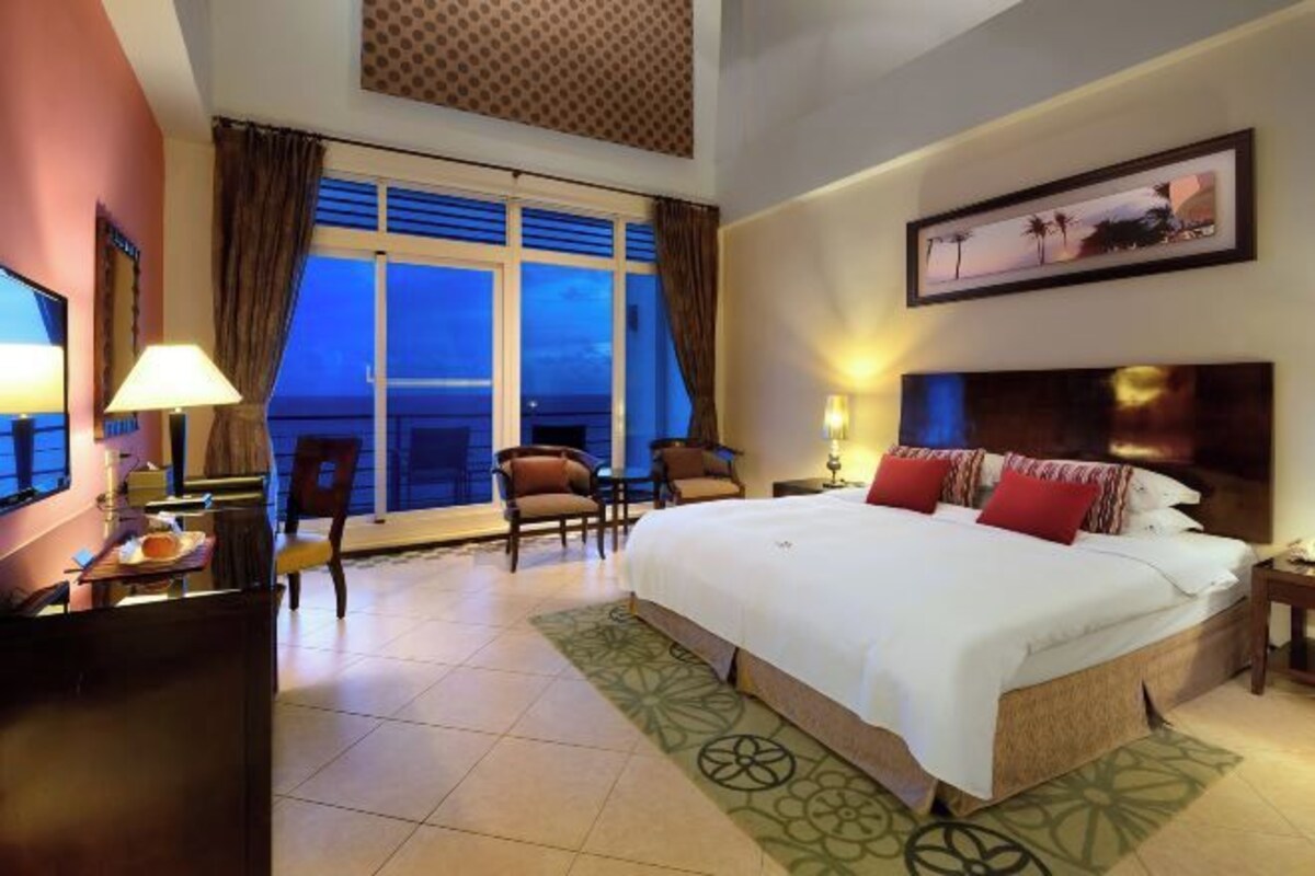 A hotel room with a large bed, a desk with a lamp, a chair, a view of the ocean, and a painting on the wall.