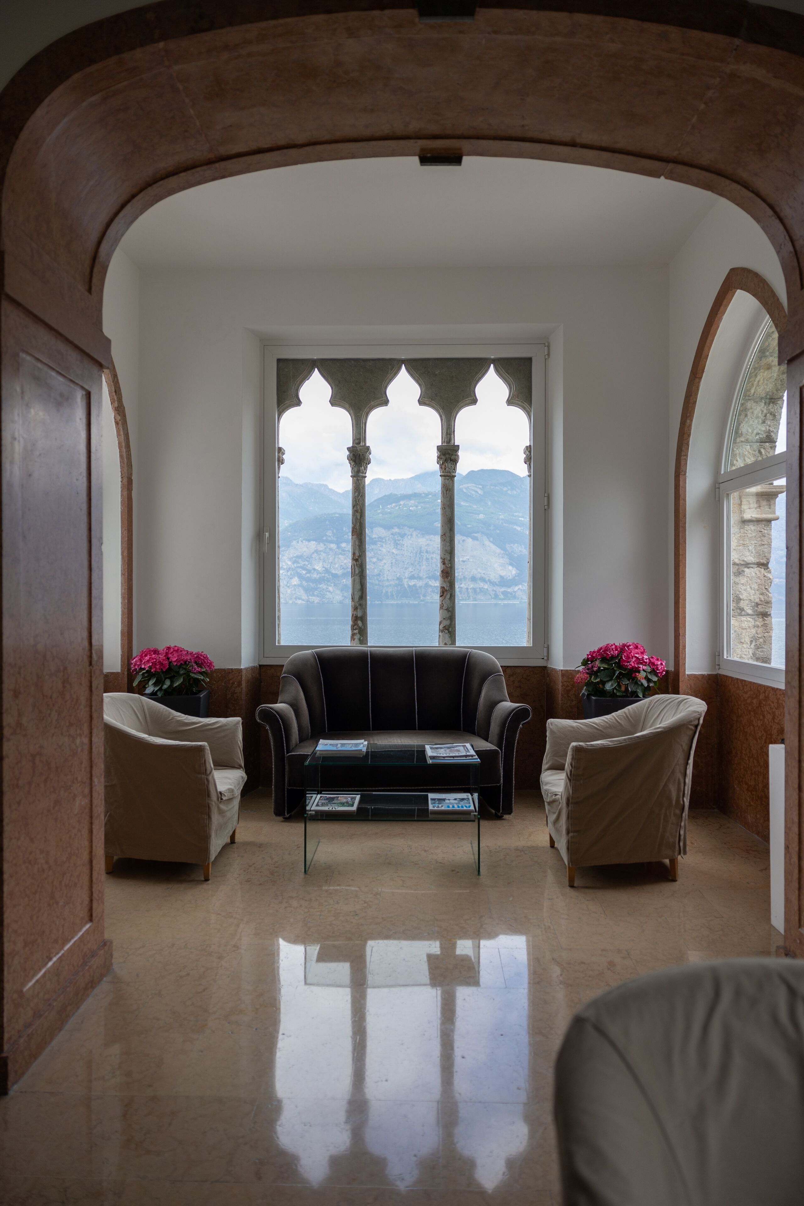 A room with a view of mountains through a window with a decorative frame. There is a sofa and two armchairs with a coffee table in the center.