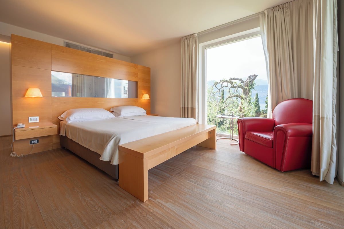 Une chambre d’hôtel moderne avec un lit en bois, des lampes de chevet, un fauteuil rouge et une grande fenêtre donnant sur un beau paysage.