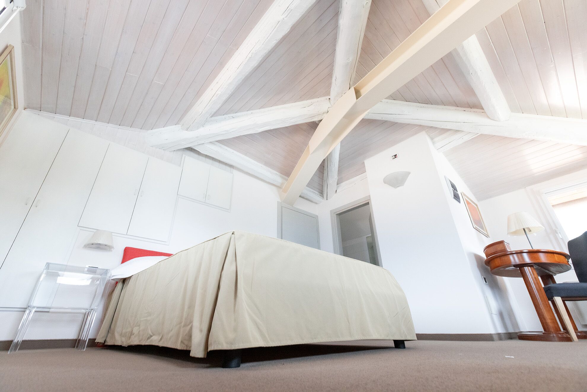Una habitación con techo alto, paredes blancas, una cama con cubierta color crema, una silla de plástico transparente y una mesa de madera.