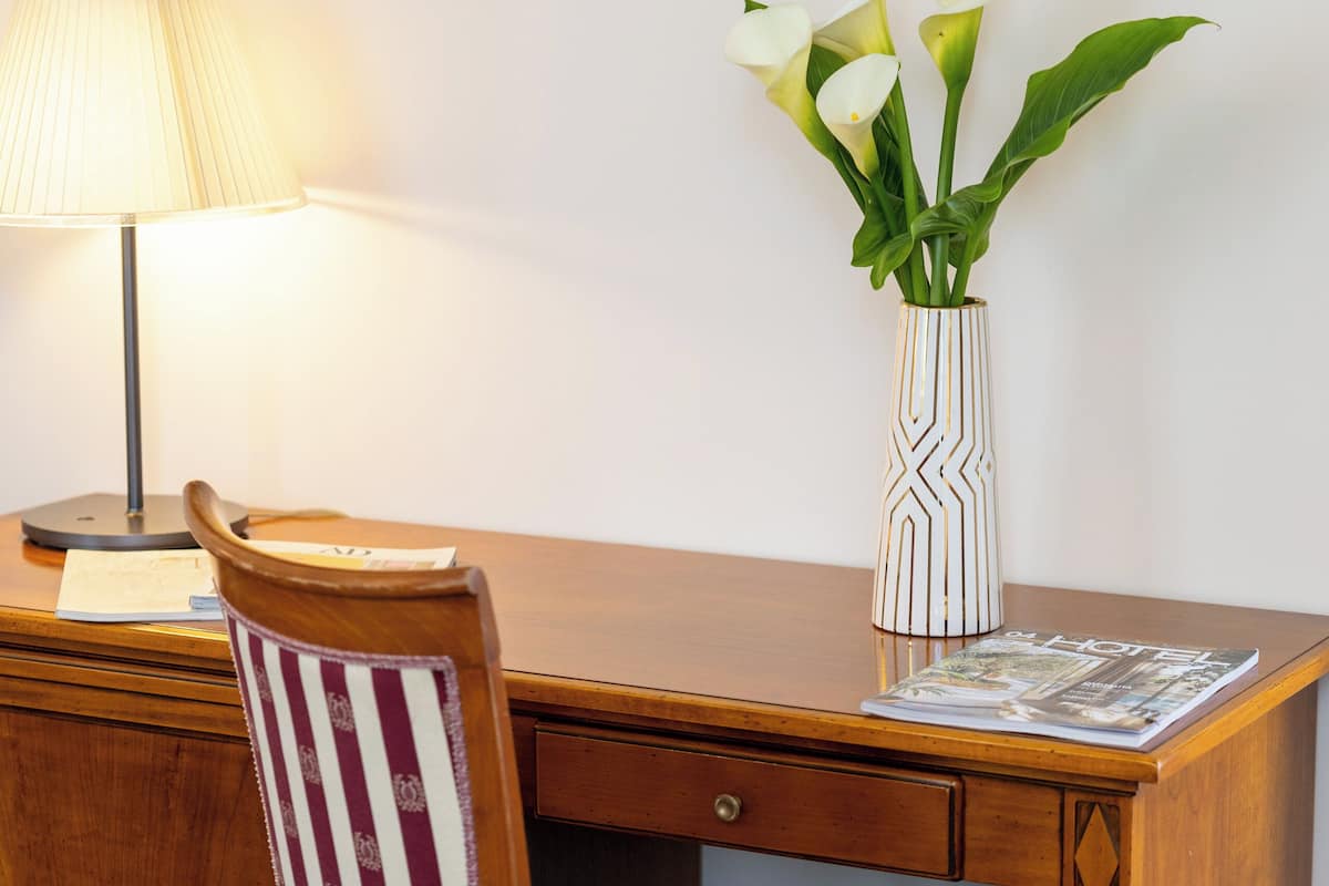 Un bureau en bois avec une lampe, une chaise à rayures, un vase de fleurs et des œuvres d’art encadrées accrochées au mur.