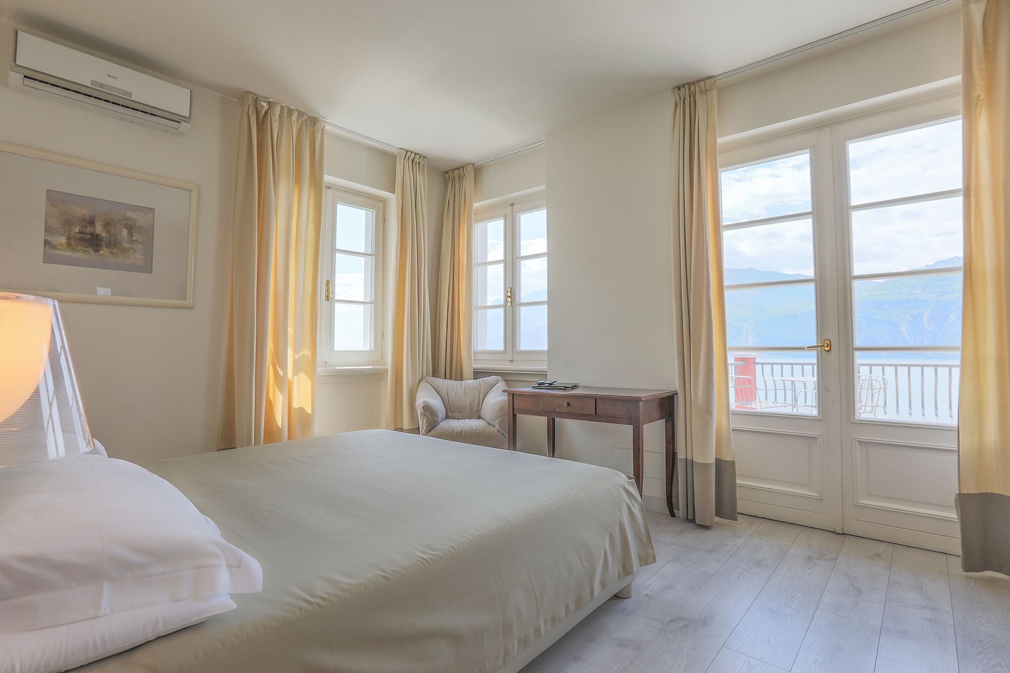 Hotelkamer met een bed, bureau en stoel. Er zijn ramen met gordijnen, en een schilderij aan de muur.