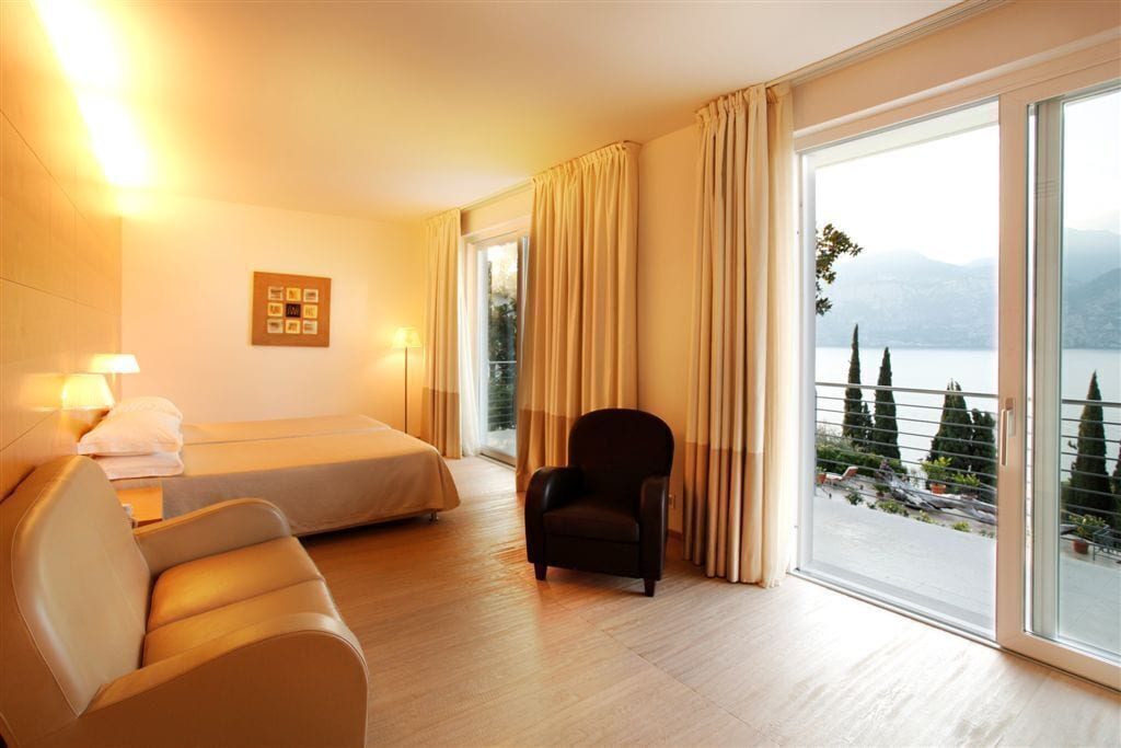 A hotel room with a bed, a chair, a sofa, and a large window with a view of trees and mountains.