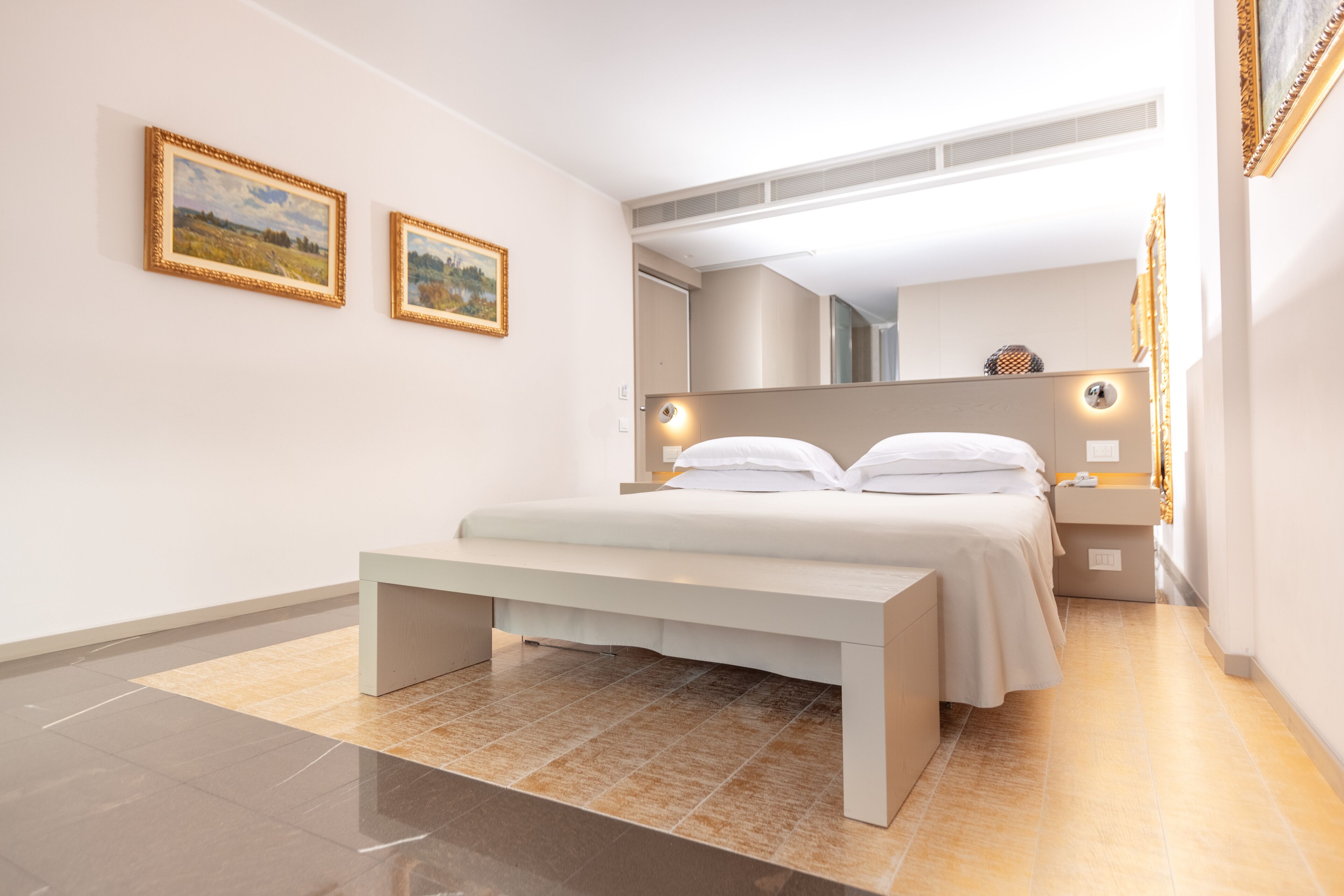 Ein modernes Schlafzimmer mit einem großen Bett, einer Bank, zwei gerahmten Bildern an der Wand und einem Spiegel über dem Bett.