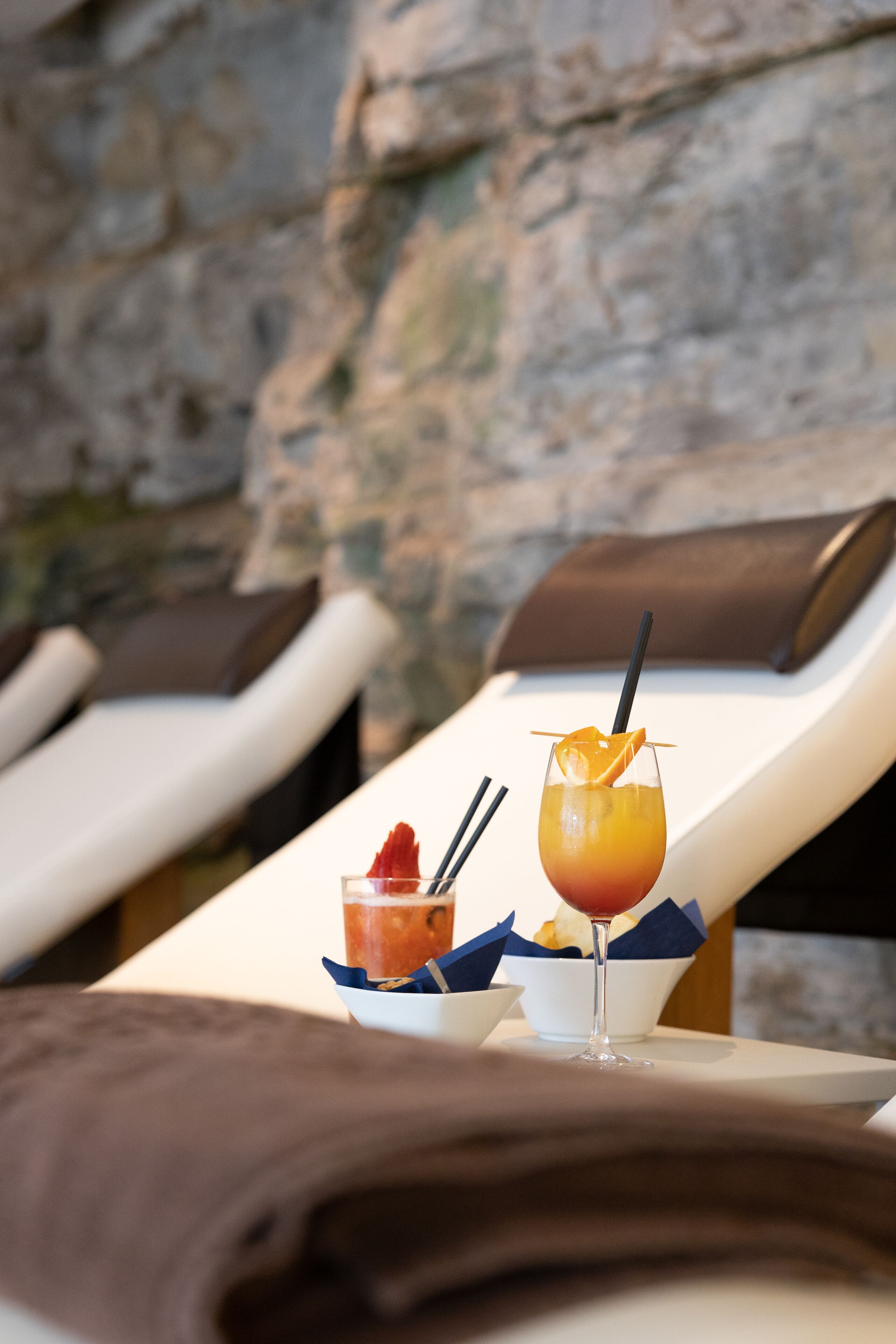 A table with a drink and a small dish, set against a stone wall with lounge chairs.