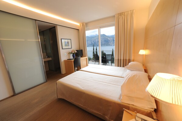 Habitación de hotel con dos camas, un escritorio con lámpara, televisión y vista a las montañas y al agua.