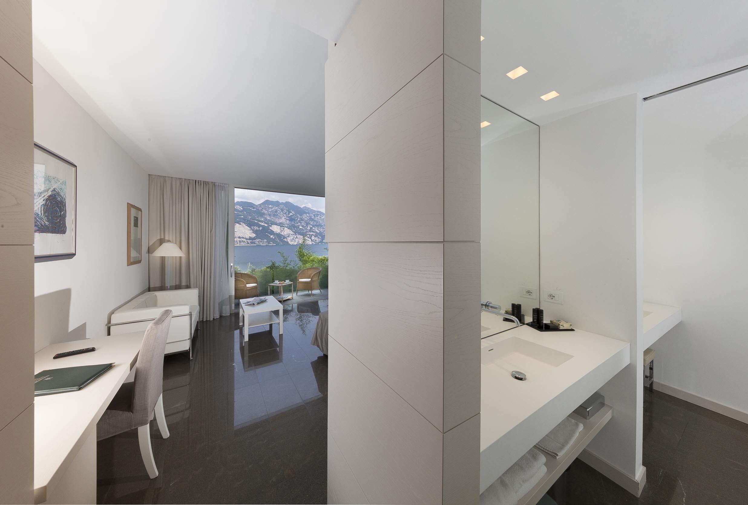 A modern hotel room with a large window offering a mountain view, a desk with a chair, a bathroom with a large mirror and a double sink.