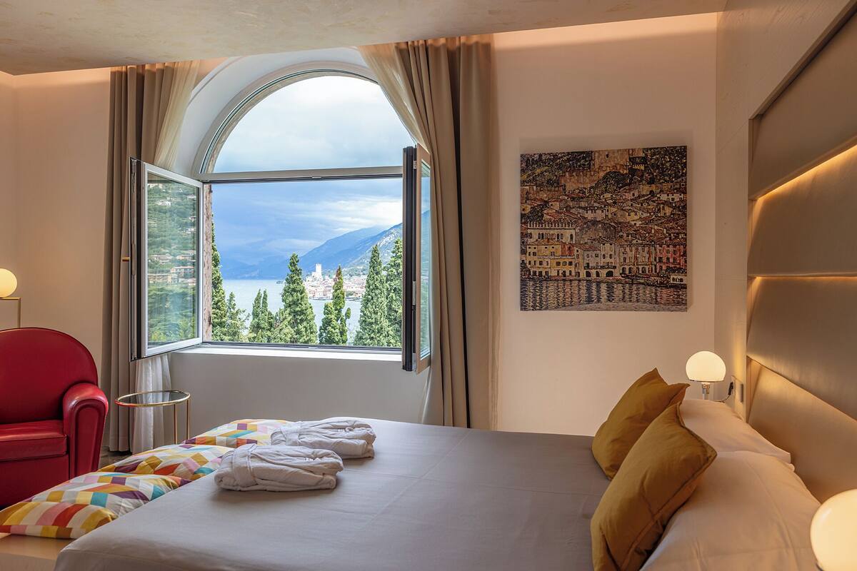 Une chambre d’hôtel avec une grande fenêtre, un lit avec des serviettes, une chaise rouge et un tableau au mur.