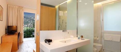 A modern bathroom with a large mirror, white sink, and a shower. There is a small bench and a television in the adjacent room.