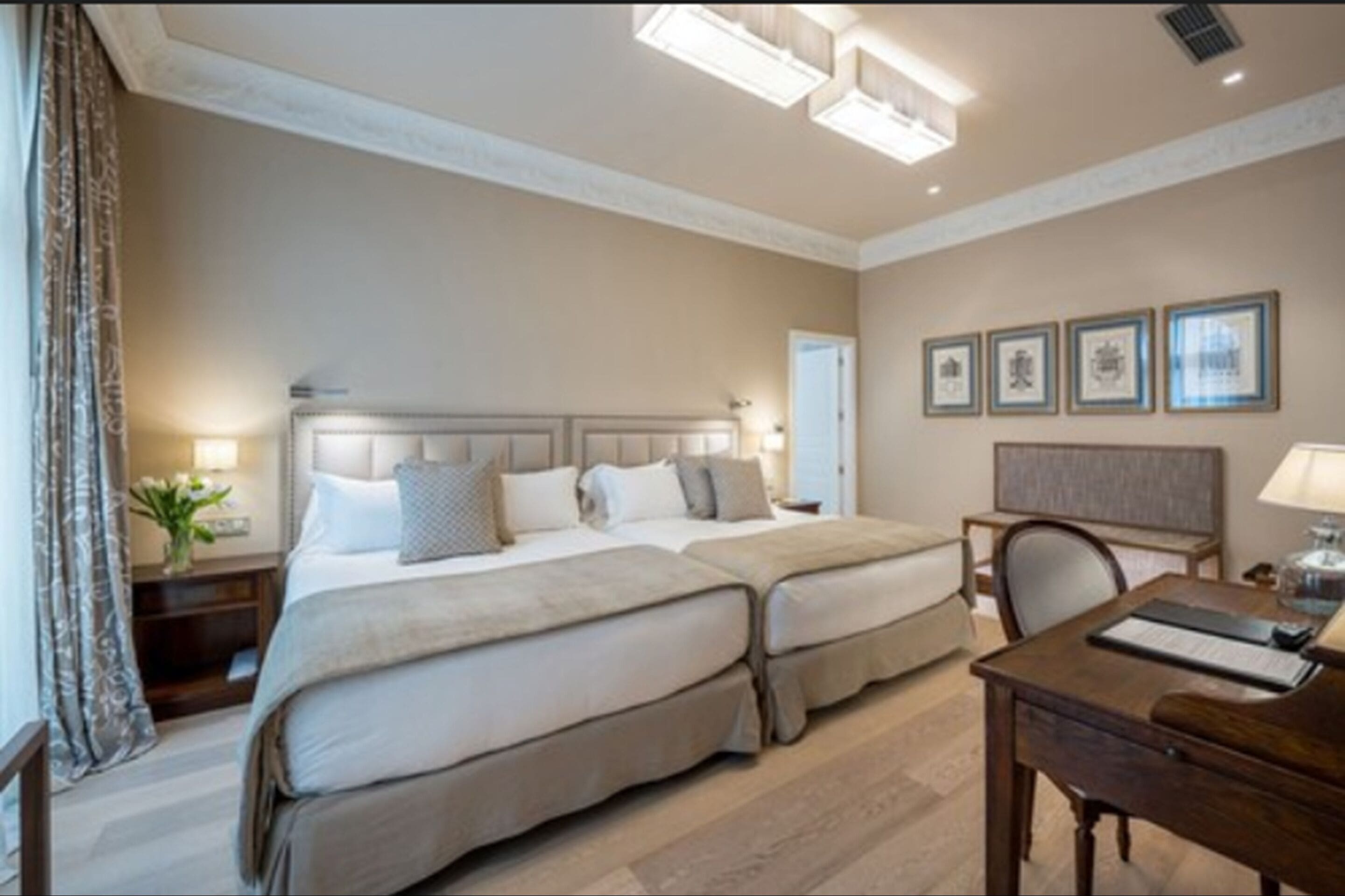 A hotel room with two beds, a desk, and a chair. There are framed pictures on the wall and a lamp on the desk.