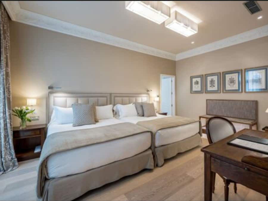 A hotel room with two beds, a desk, and a chair. There are framed pictures on the wall and a lamp on the desk.