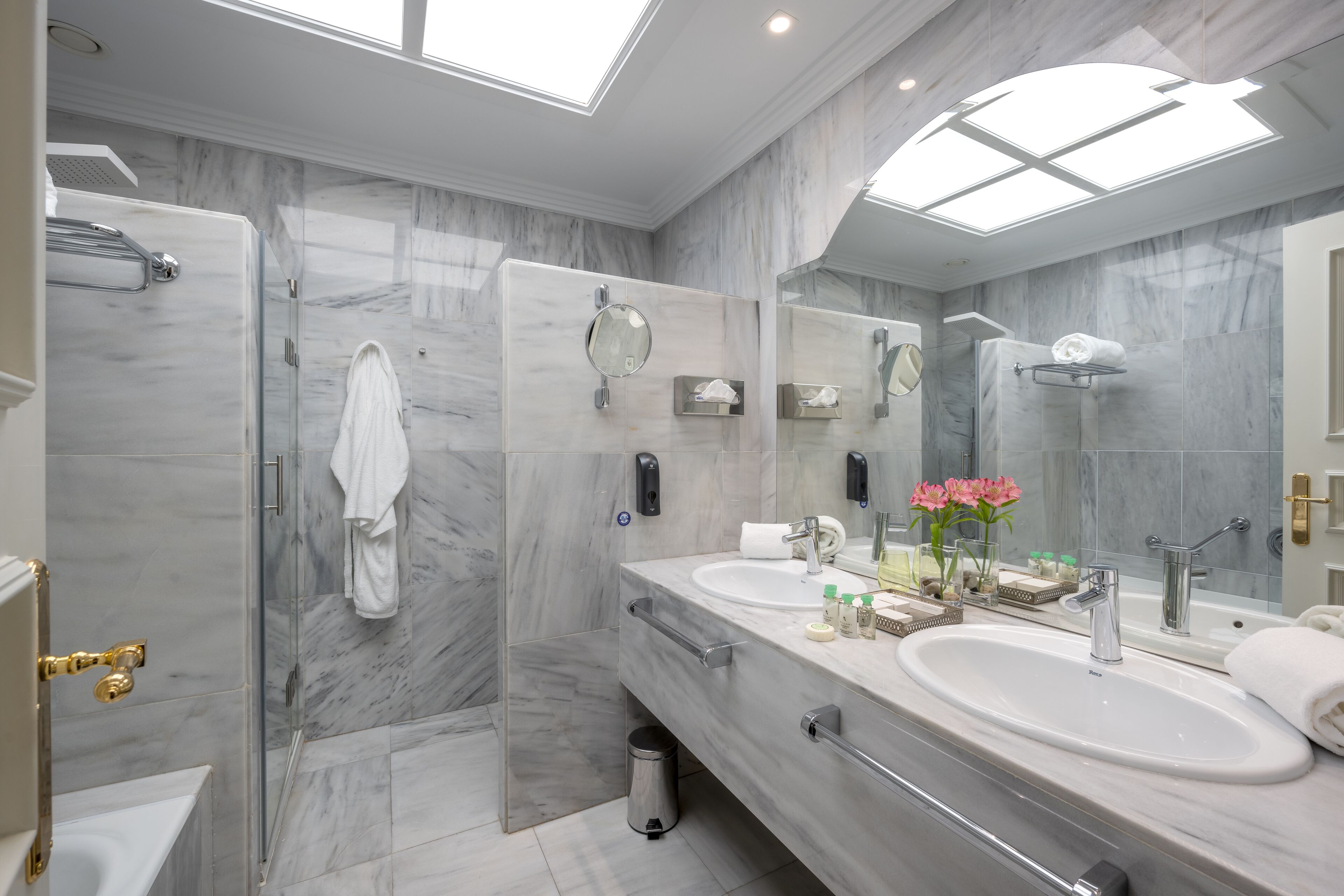 Un baño moderno con ducha envuelta en vidrio, dos lavamanos, un espejo grande y un tragaluz.