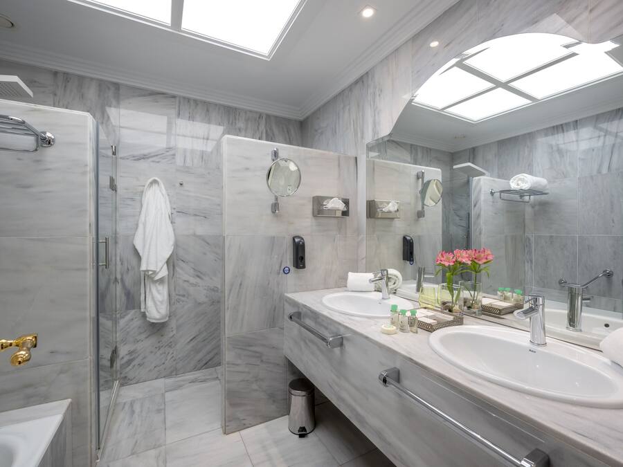 Un baño moderno con ducha envuelta en vidrio, dos lavamanos, un espejo grande y un tragaluz.