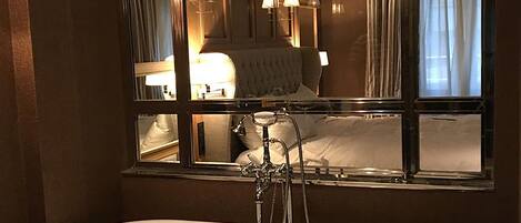 Un baño con un espejo grande, una bañera y una cama visible en el reflejo.