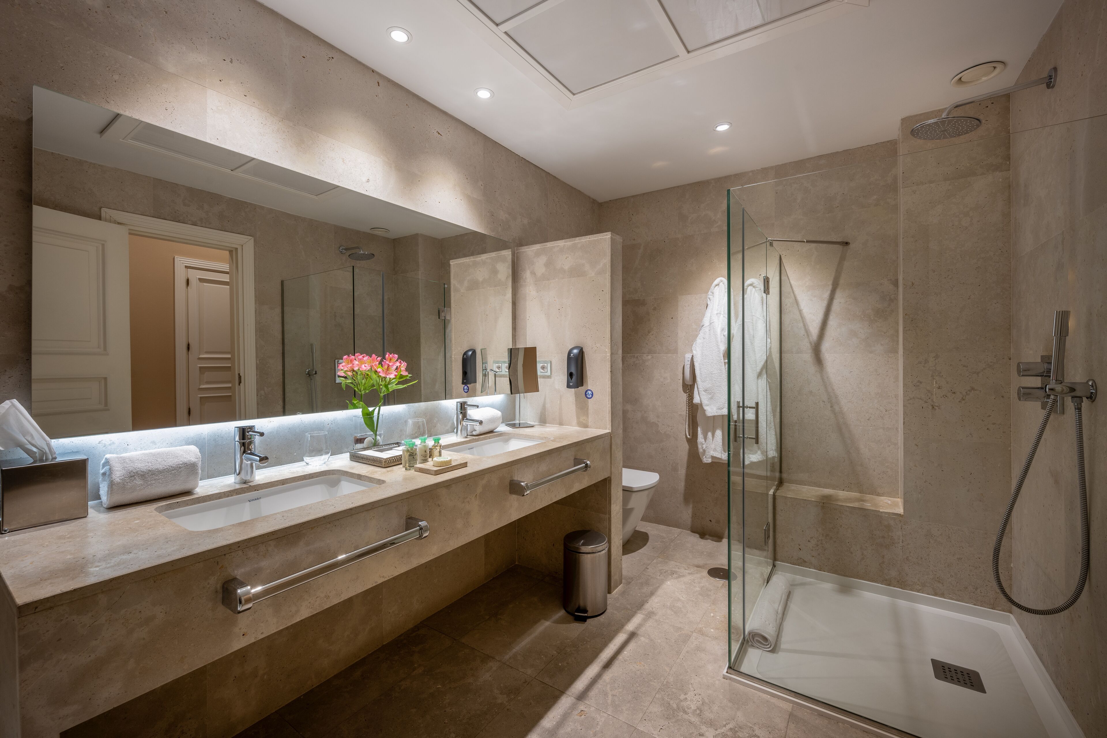A modern bathroom with a large mirror, two sinks, a glass shower enclosure, and a toilet.