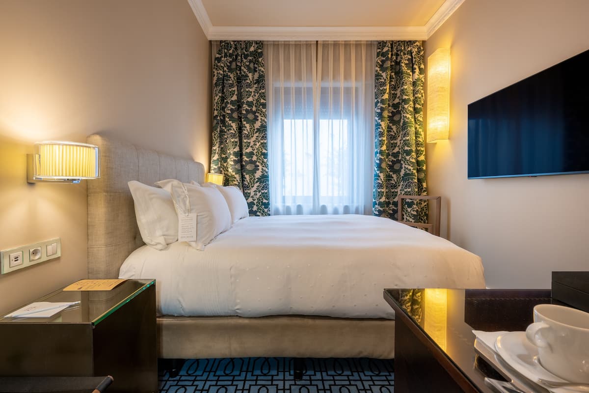 A hotel room with a bed, bedside lamps, a flat-screen TV, a desk with a cup and saucer, and patterned curtains.