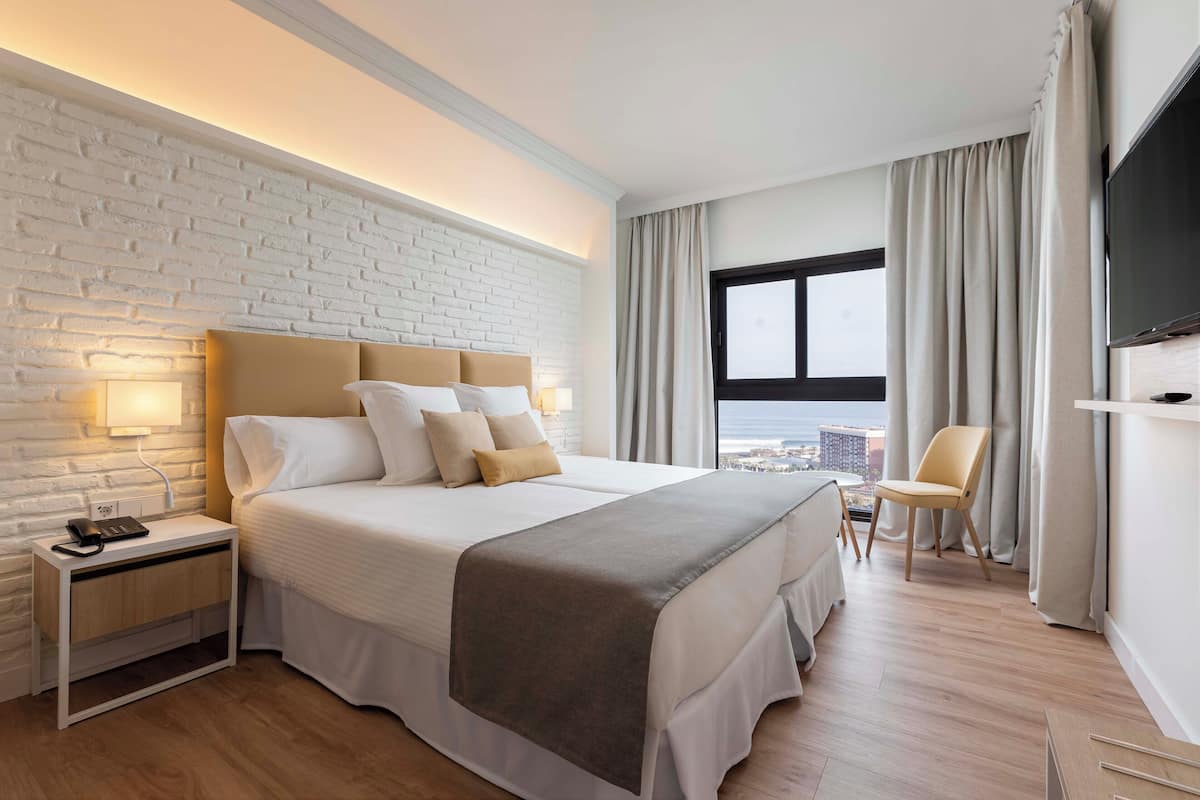 A modern hotel room with a large bed, a nightstand, a TV, and a view of the city.