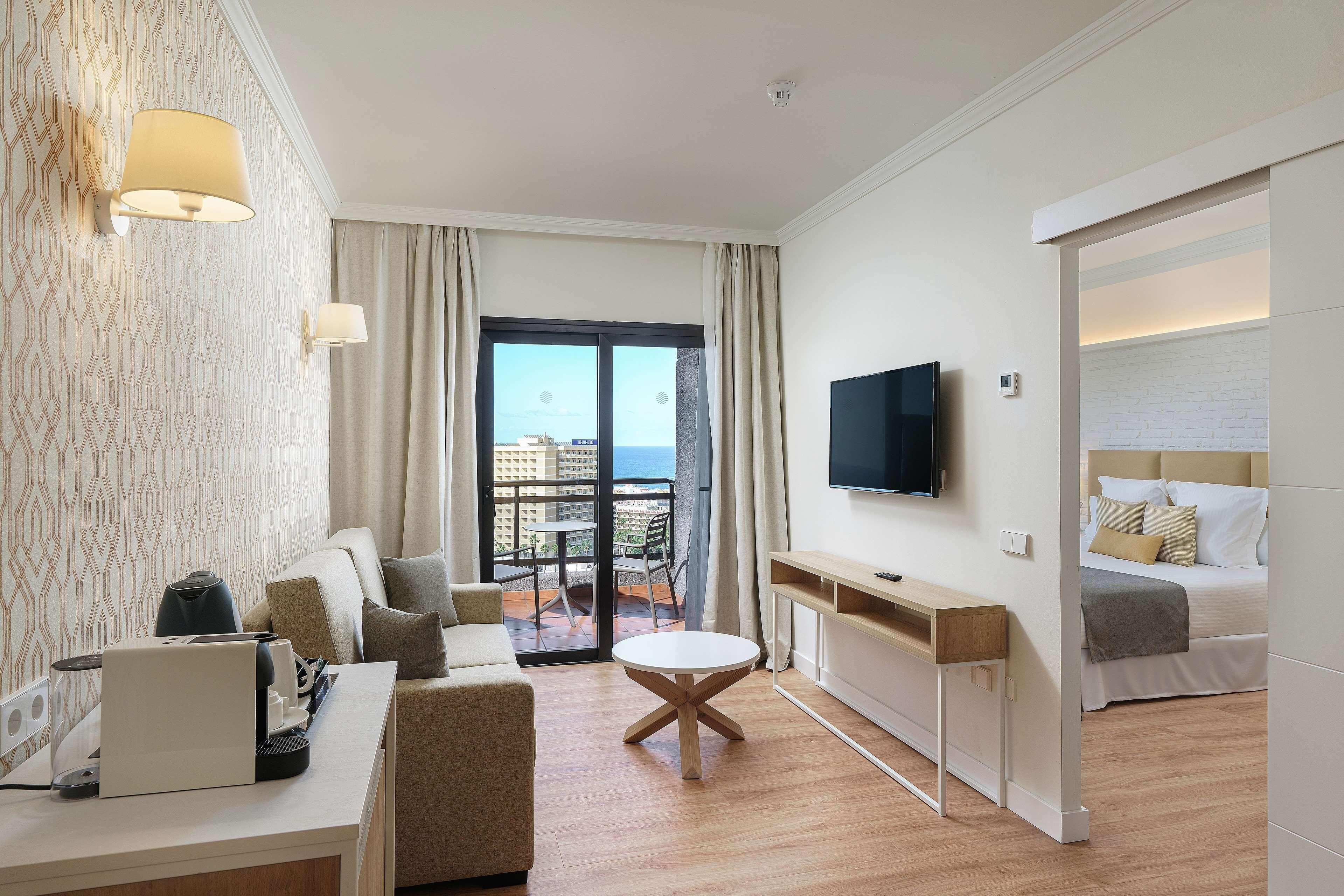 A hotel room with a sofa, a round table, a TV, and a balcony with a view.