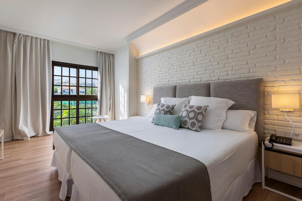 A modern hotel room with a large bed, a white brick accent wall, a nightstand with a phone, and a view of greenery through the window.