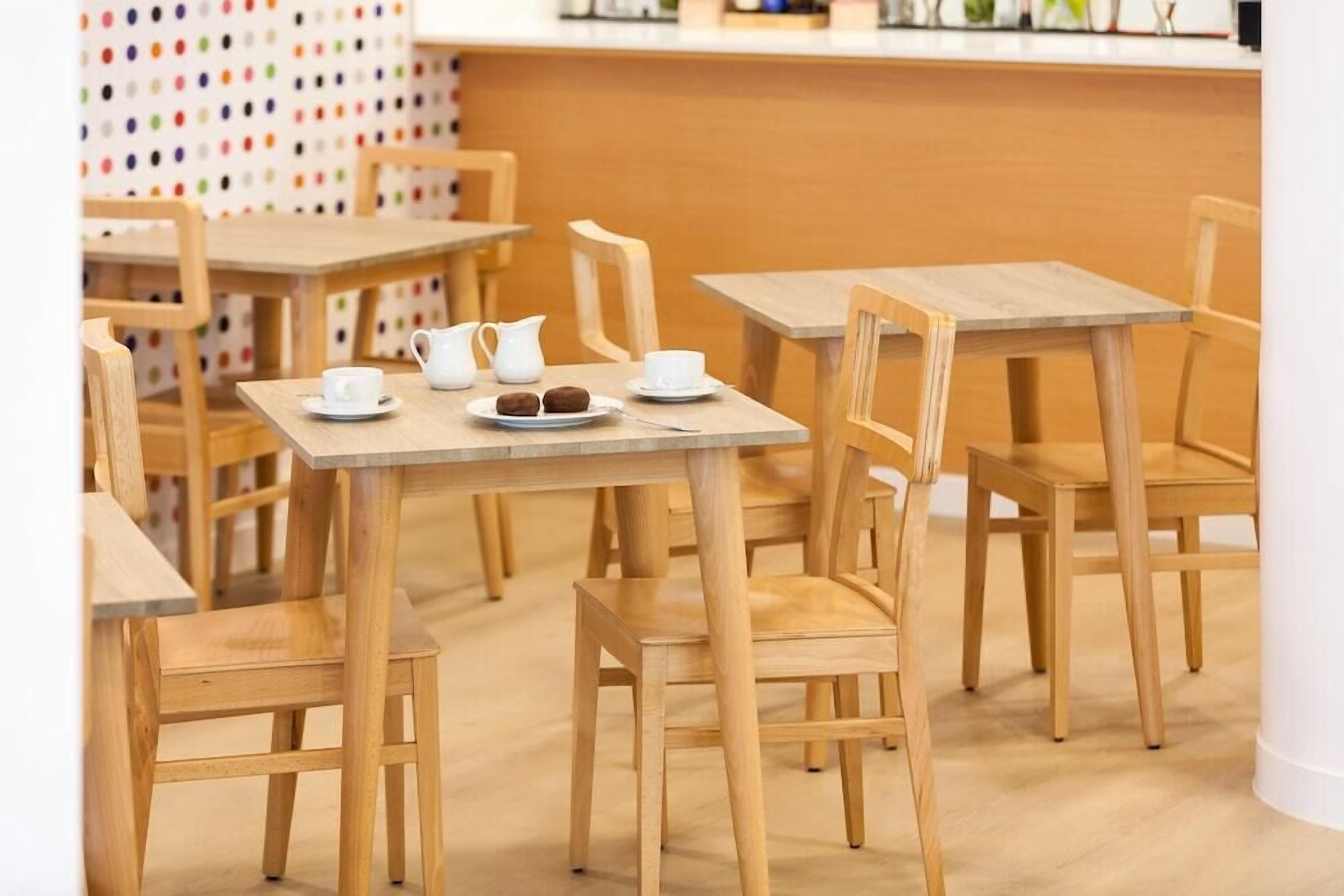 A café with wooden tables and chairs, a white pitcher, cups, and a plate with pastries.