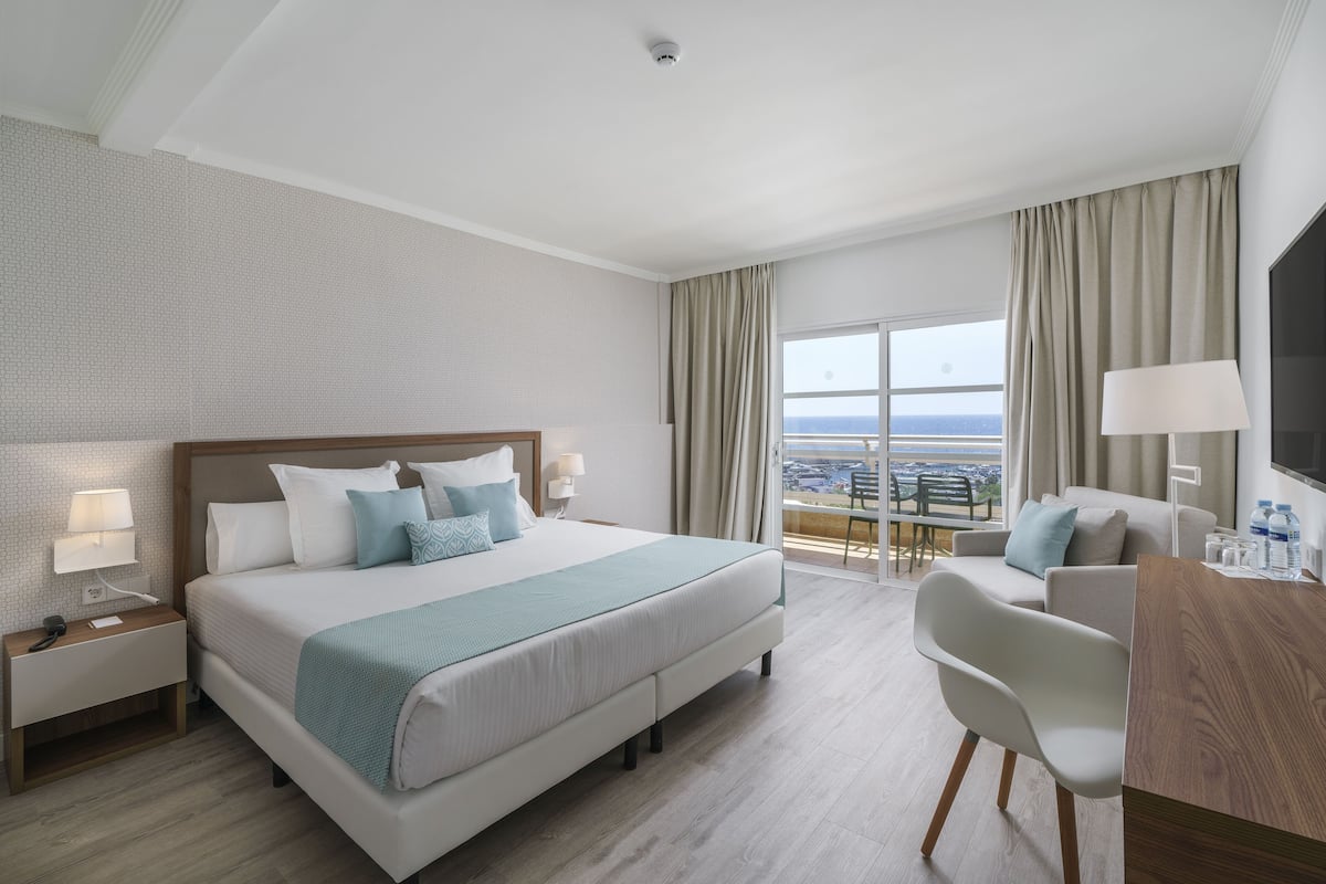 A hotel room with a large bed, a desk, a chair, a TV, and a balcony with a view.