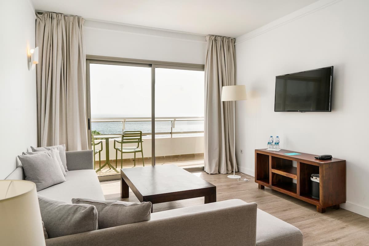 A modern hotel room with a large window, a sofa, a coffee table, a TV, and a wooden sideboard.