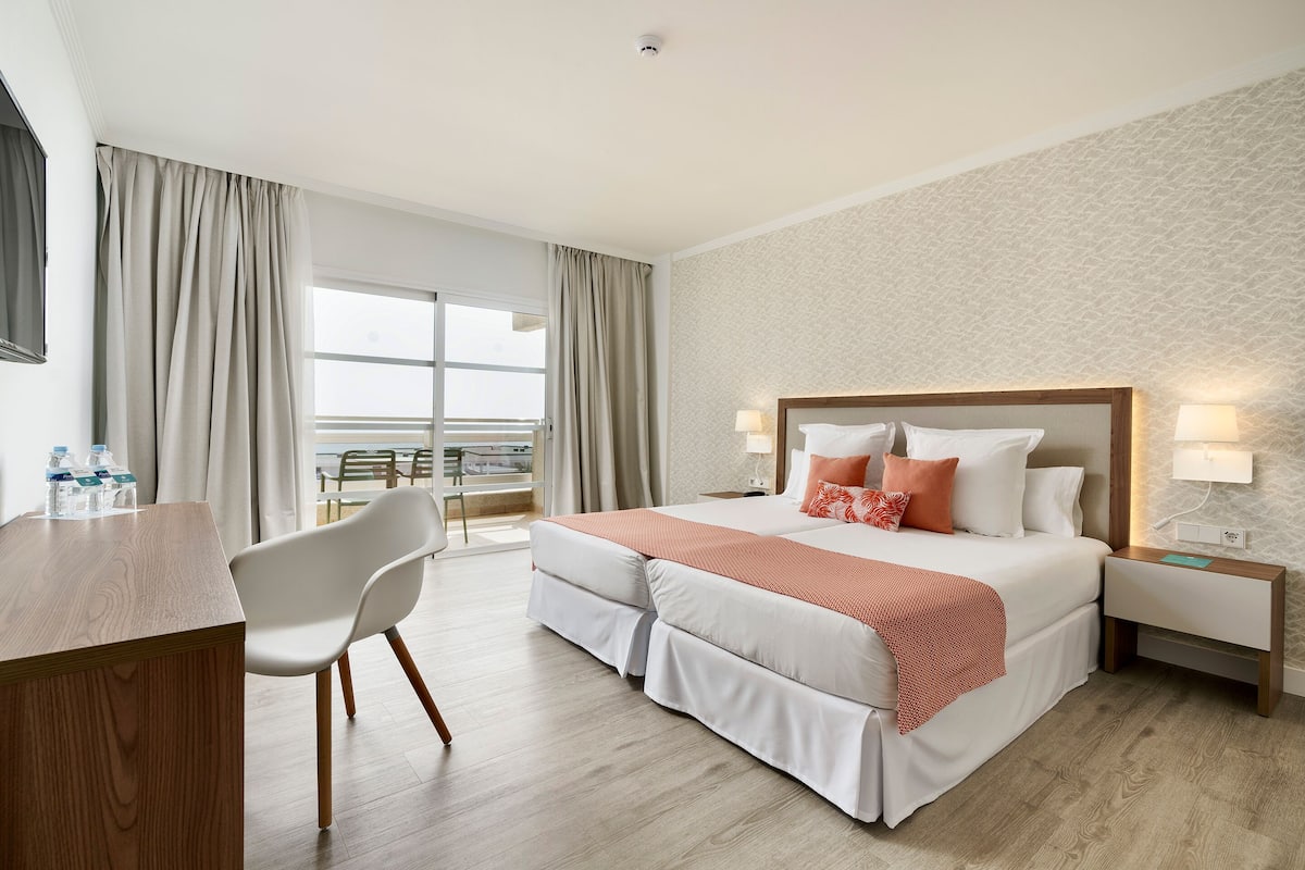 A hotel room with a bed, bedside tables, a desk, and a chair. There is a view of the outdoors through a large window.