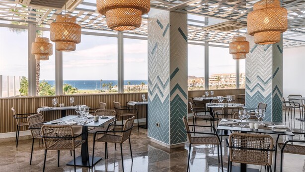 A restaurant with outdoor seating, wicker chairs, and hanging rattan lamps. Tables are set with wine glasses and plates. - Alua Atlantico Golf (San Miguel de Abona)