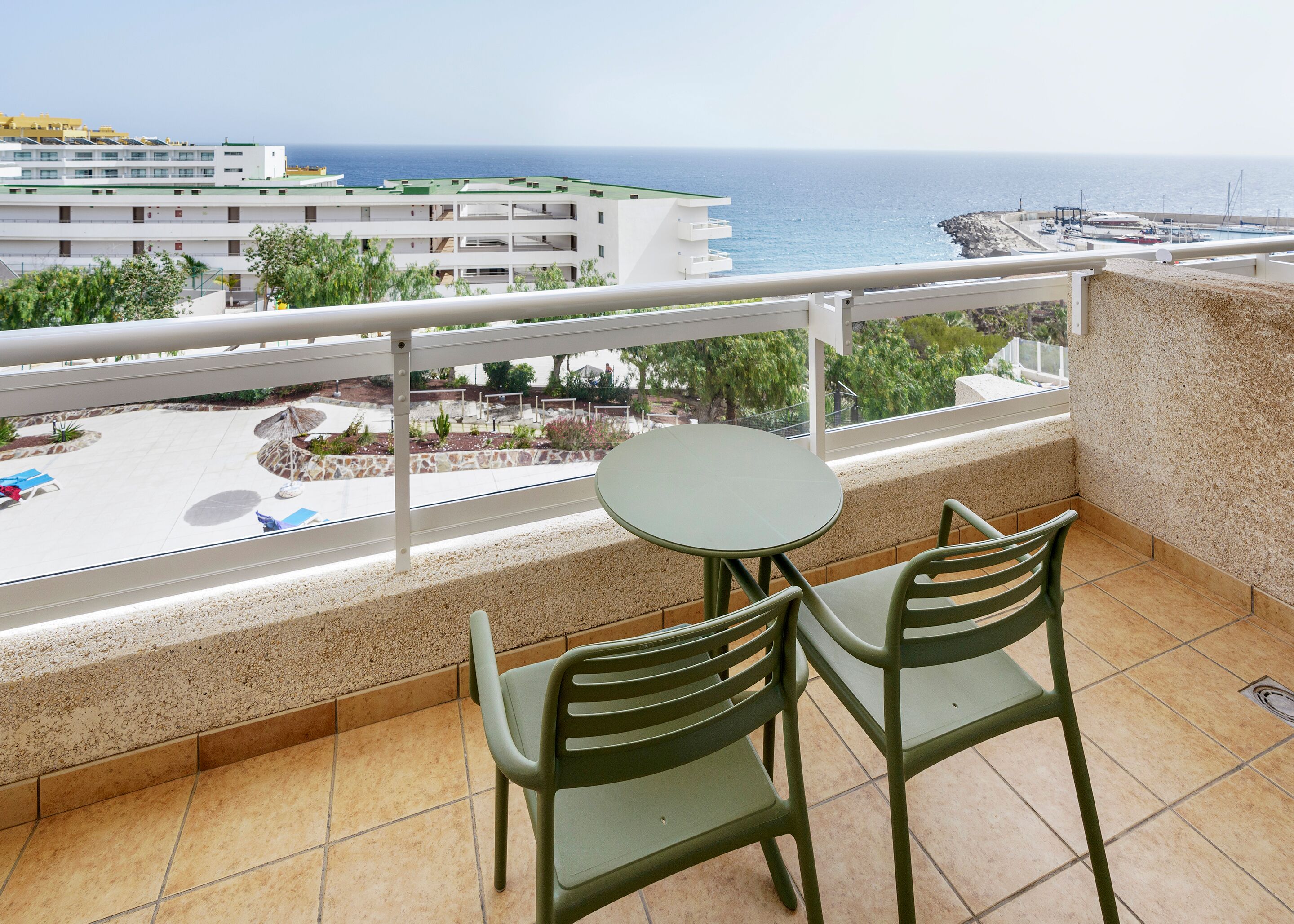 A balcony with two chairs and a table, overlooking a beach with a marina and buildings in the background.