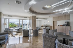 A modern hotel lobby with a spacious seating area, a kitchen, and large windows offering a view of the city.