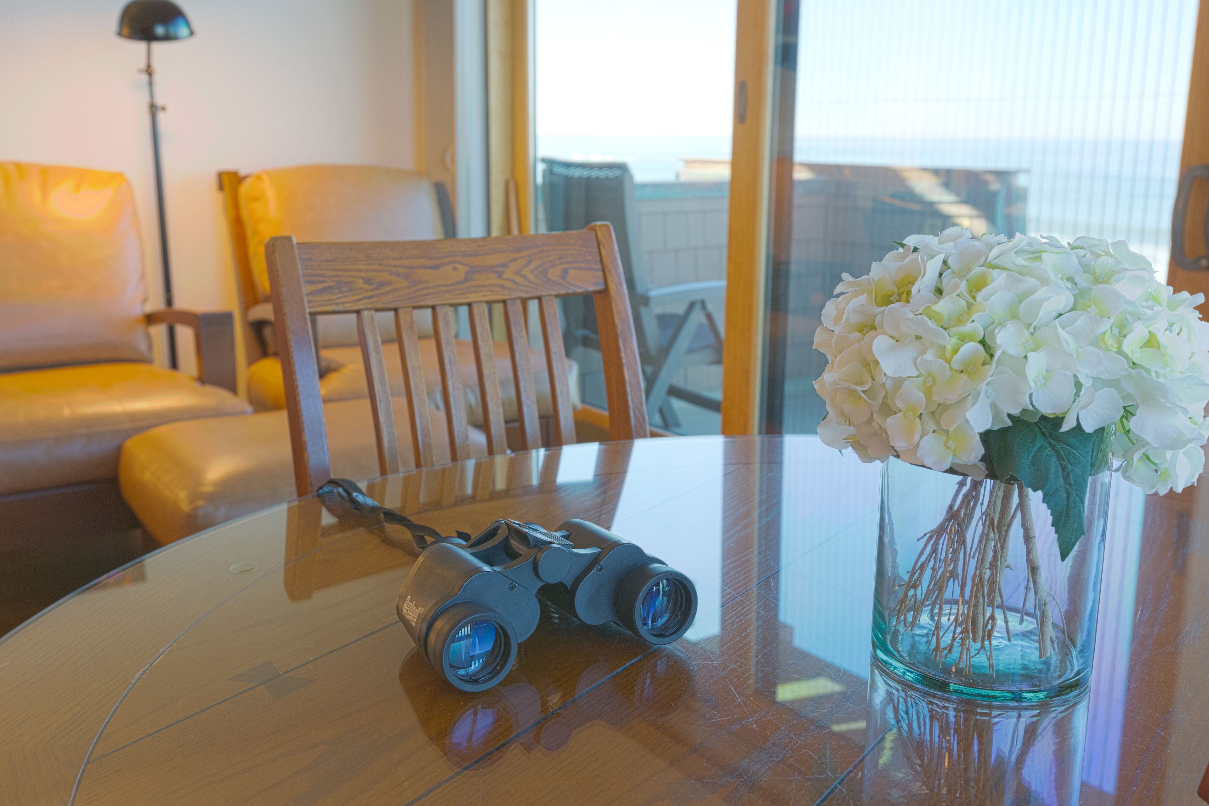 Une chaise en bois, une paire de jumelles et un vase avec des fleurs blanches sur une table en verre.