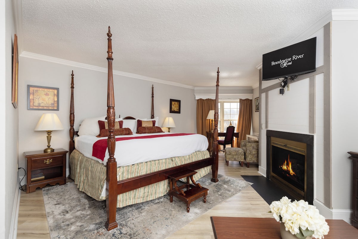 A four-poster bed with a red throw blanket, a fireplace, and a television displaying the hotel name.