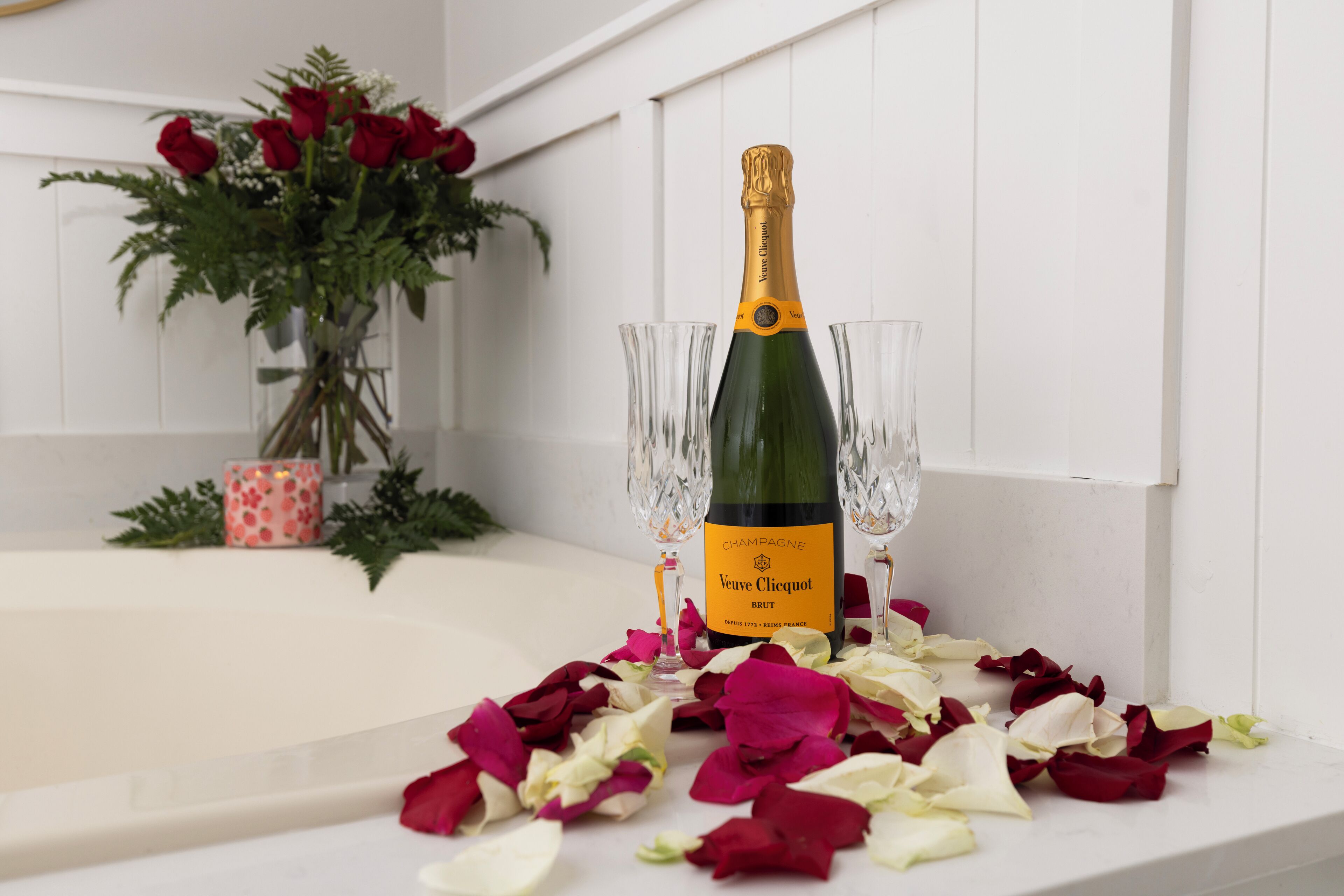 A bottle of Veuve Clicquot Brut champagne with two crystal glasses, surrounded by rose petals on a white surface.