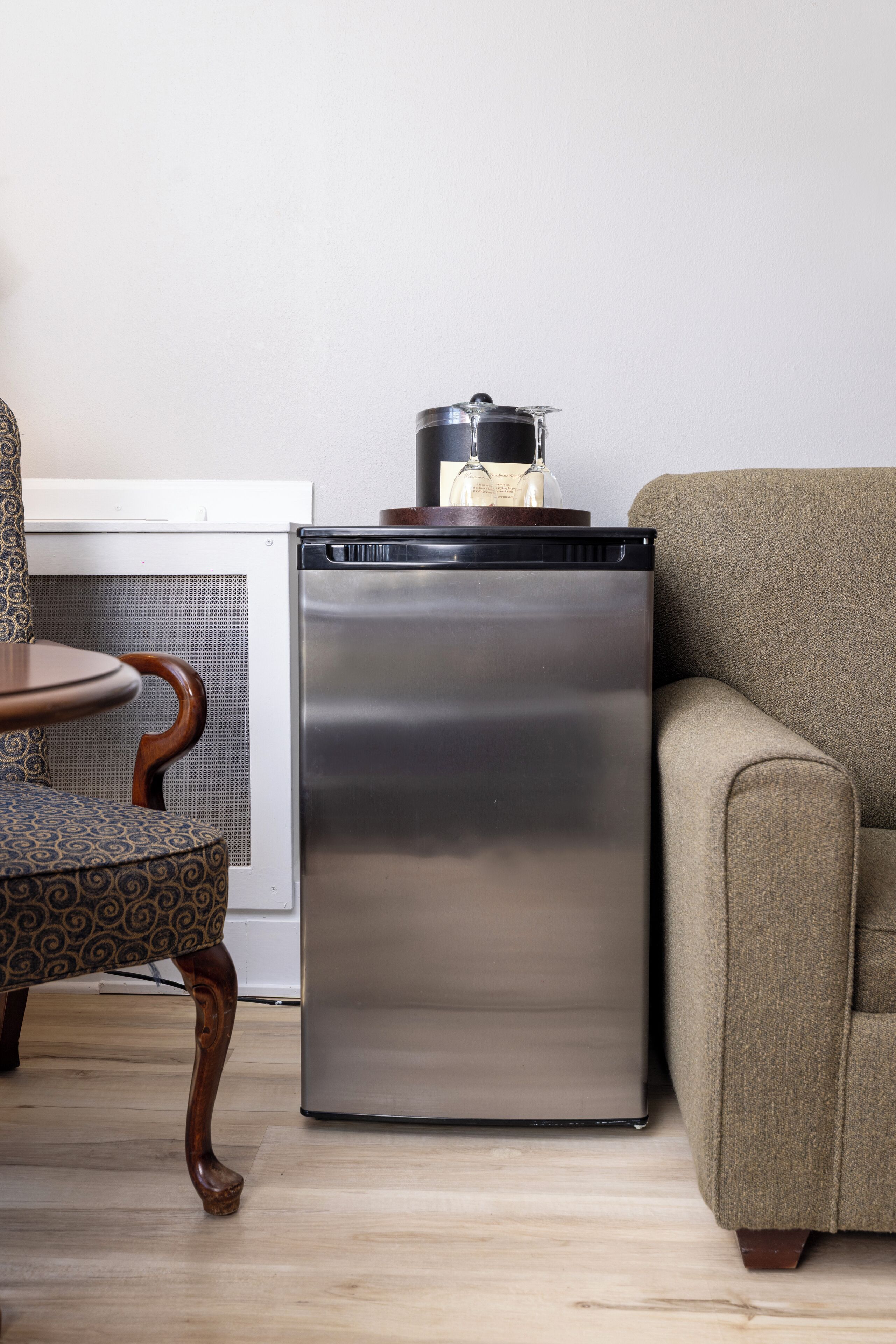 A compact refrigerator with a pot on top, a patterned chair, and a sofa in a room with a light-colored floor and plain walls.