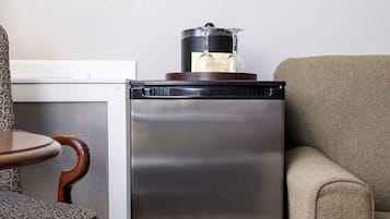 A compact refrigerator with a pot on top, a patterned chair, and a sofa in a room with a light-colored floor and plain walls.