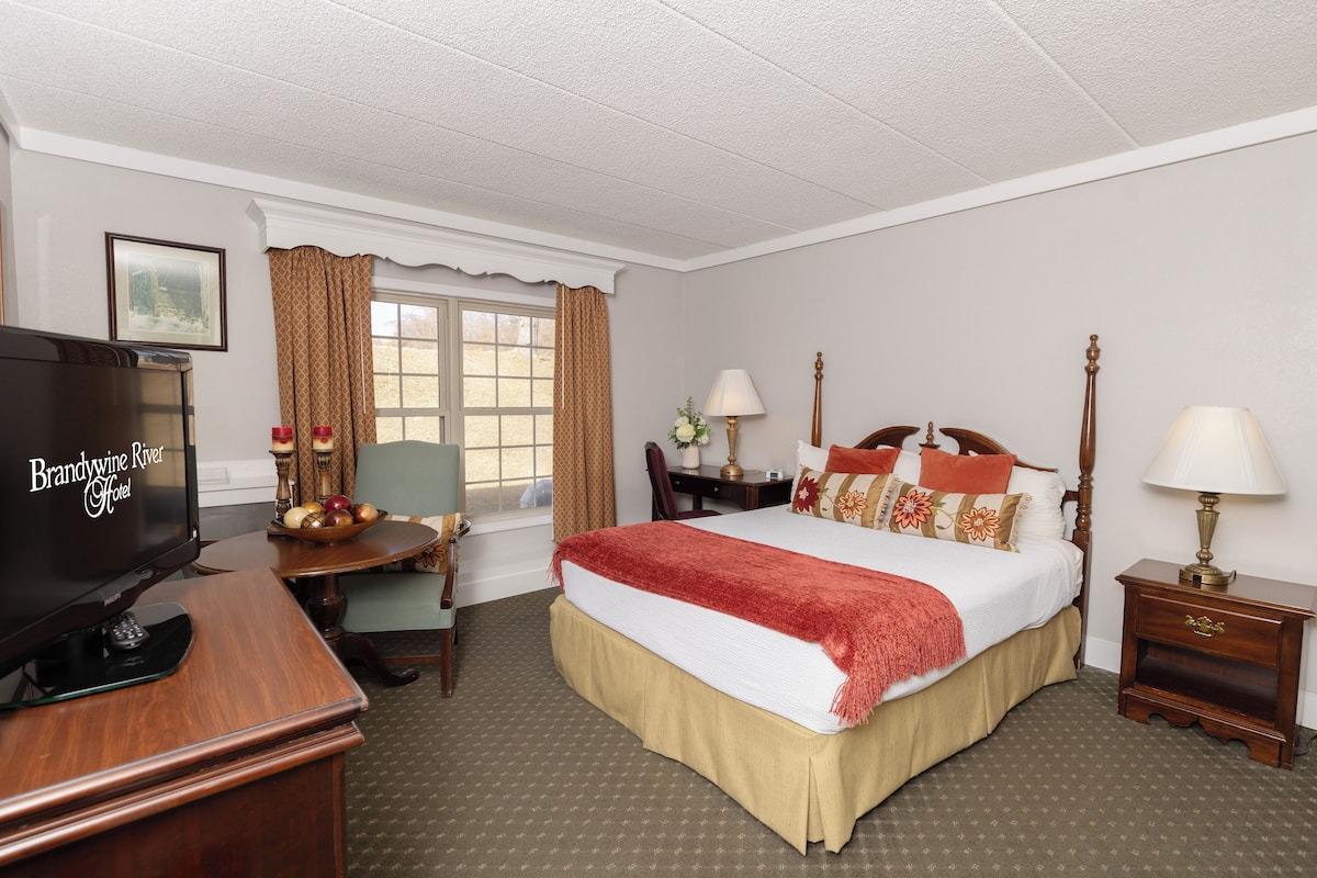 A hotel room with a bed, a desk with a television, a chair, a nightstand with a lamp, and a window with curtains.