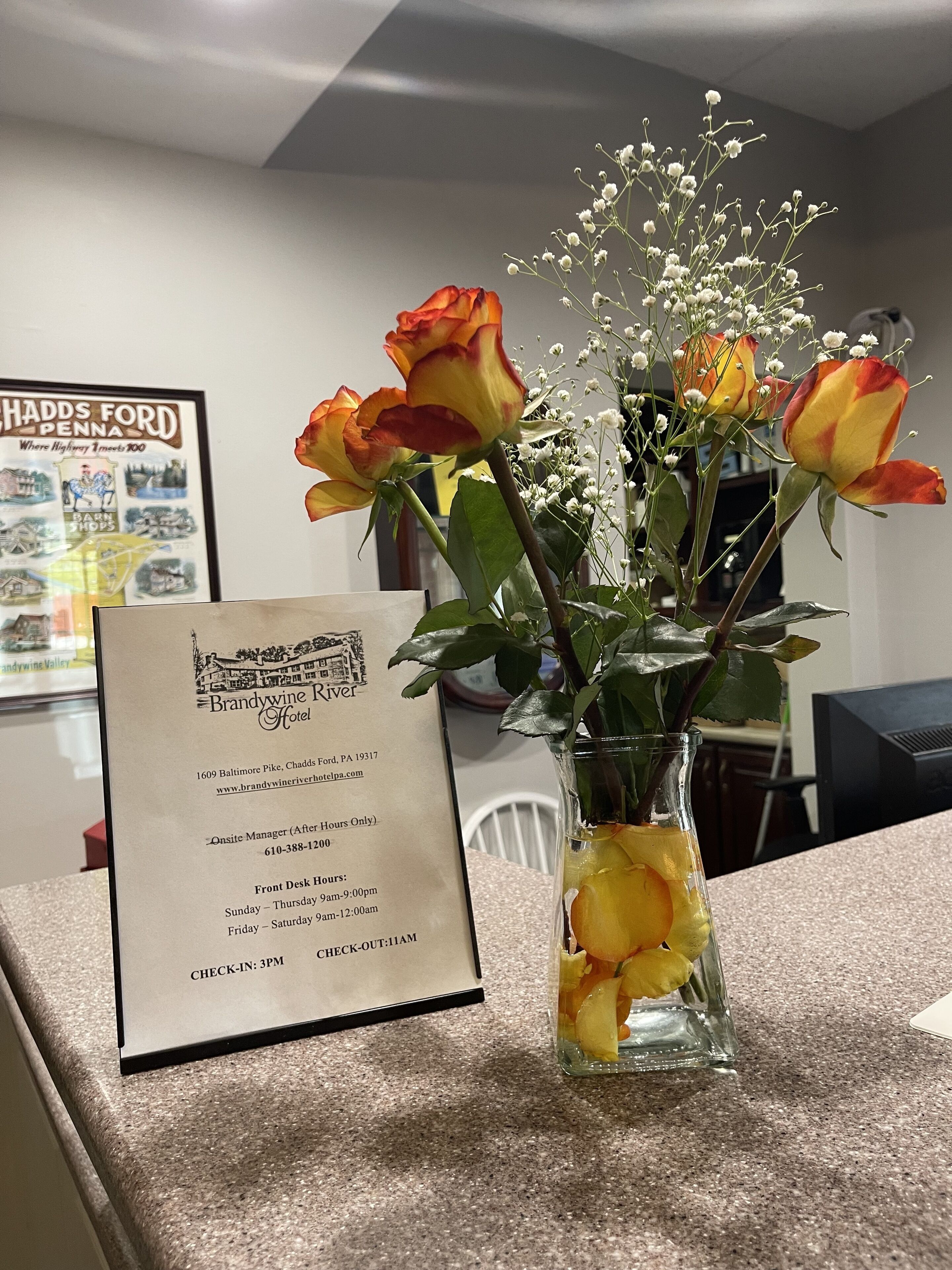 A glass vase with roses and baby's breath on a counter. A framed sign with hotel information is visible in the background.